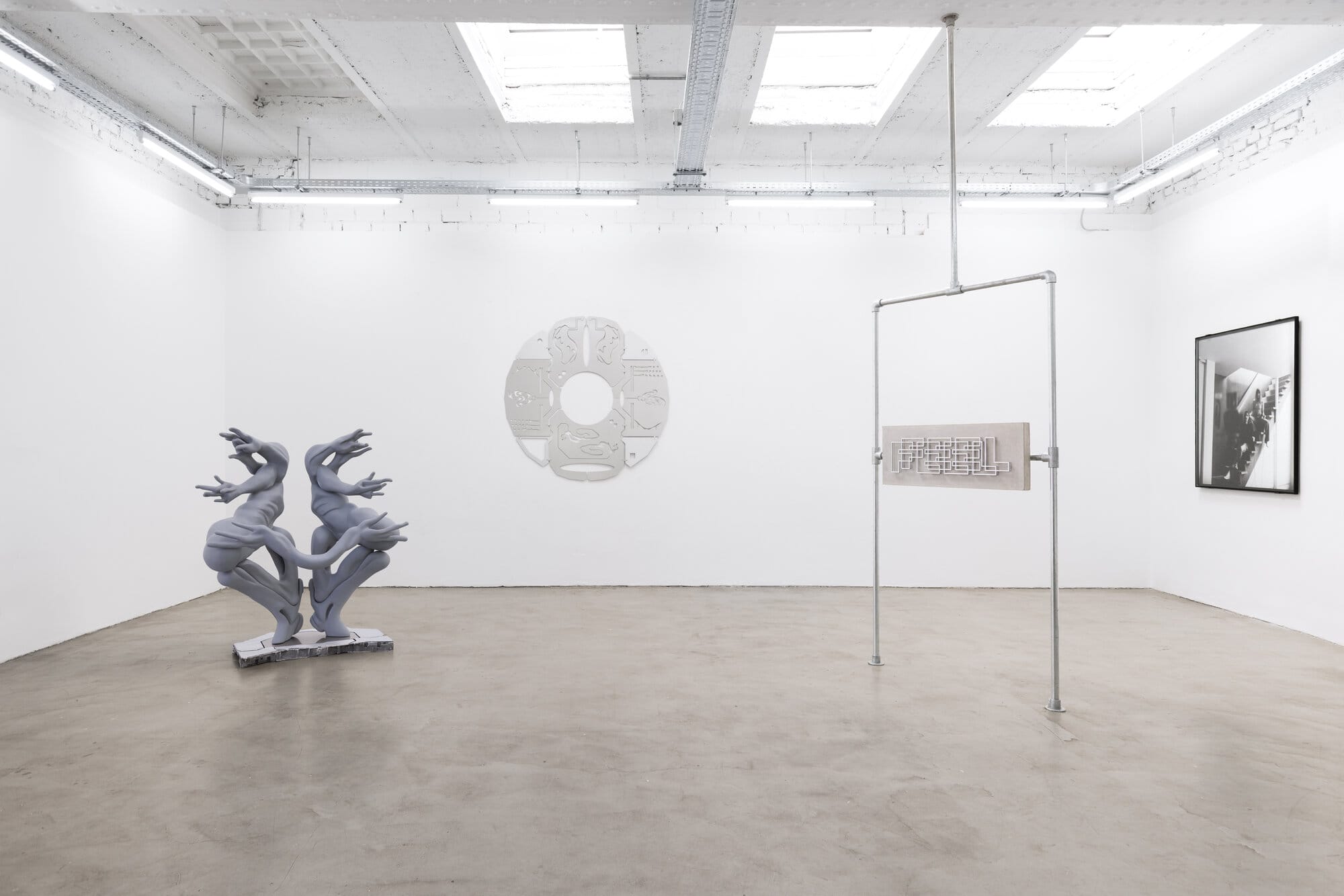 Installation view of Call Me We by lom-of-LaMa at SUPERRAUM Dortmund, 2026, showing dual-body sculpture, circular wall relief, pipe scaffold with concrete panel, and framed black-and-white photograph