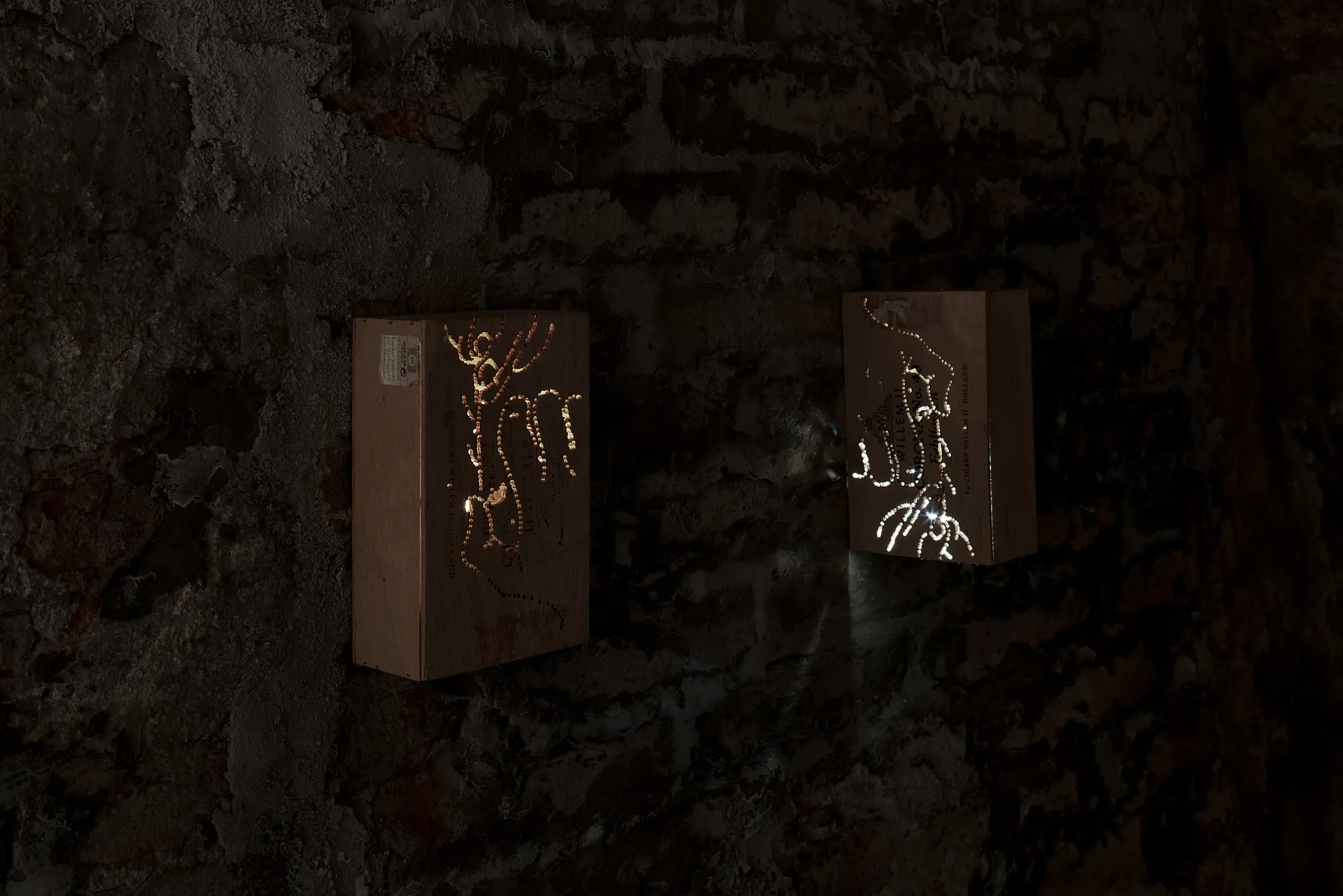 Anzhelika Palyvoda, Echoes that refuse to fade, FAVU Gallery Brno, 2026, two perforated cigar boxes mounted on dark brick wall with light through engravings