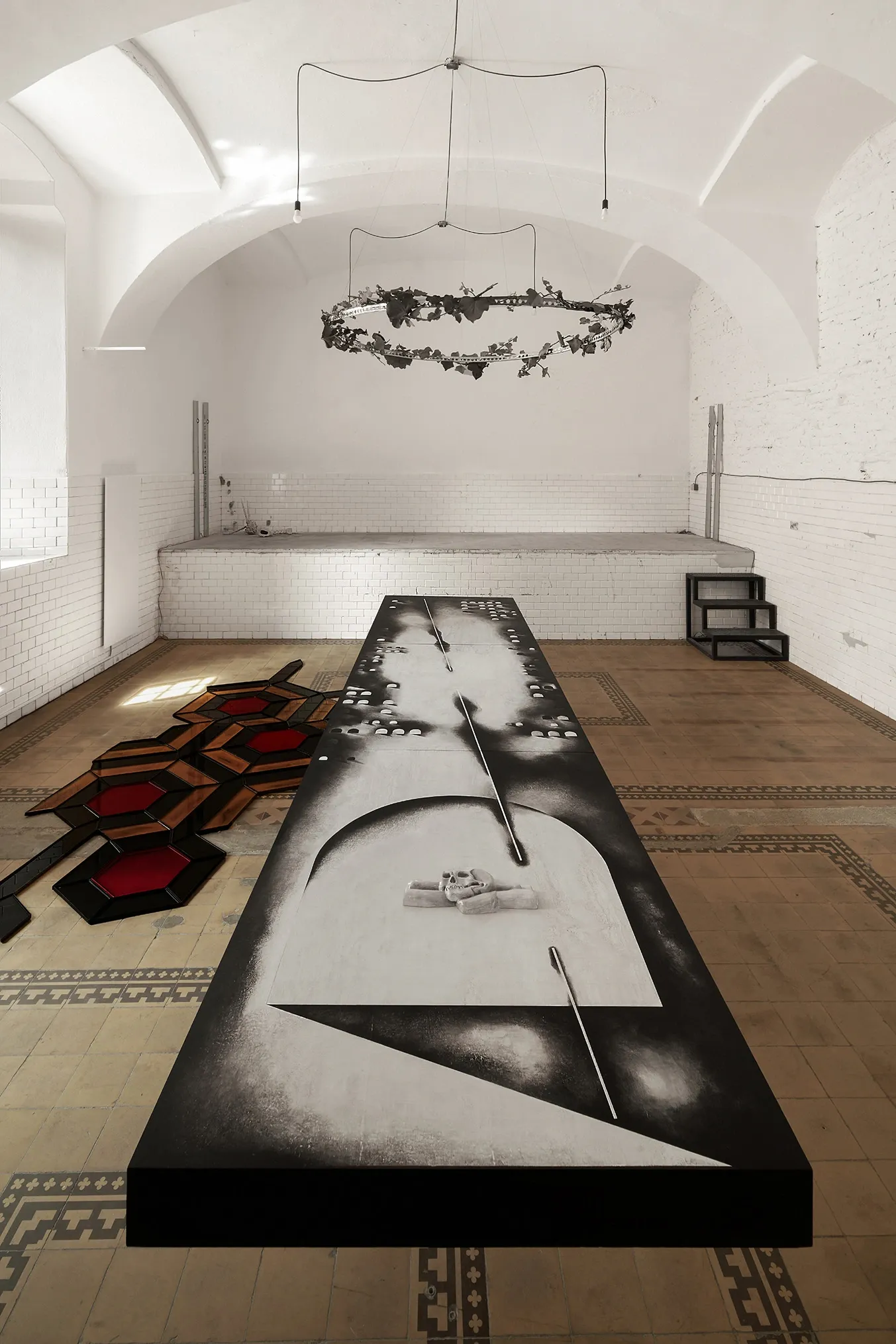 Palyvoda Struger Yesakova, Echoes that refuse to fade, FAVU Gallery Brno, 2026, wide room view with table surface skull imagery and hexagonal floor grid