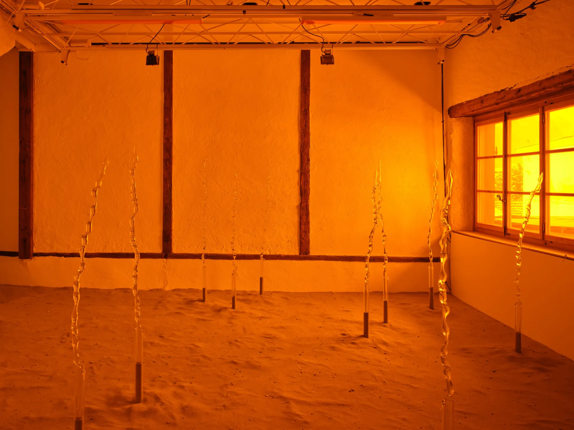 Rahel Oberhummer, Sentinels, 2025, wide room view with multiple glass rods and sand floor bathed in amber light, Montreux