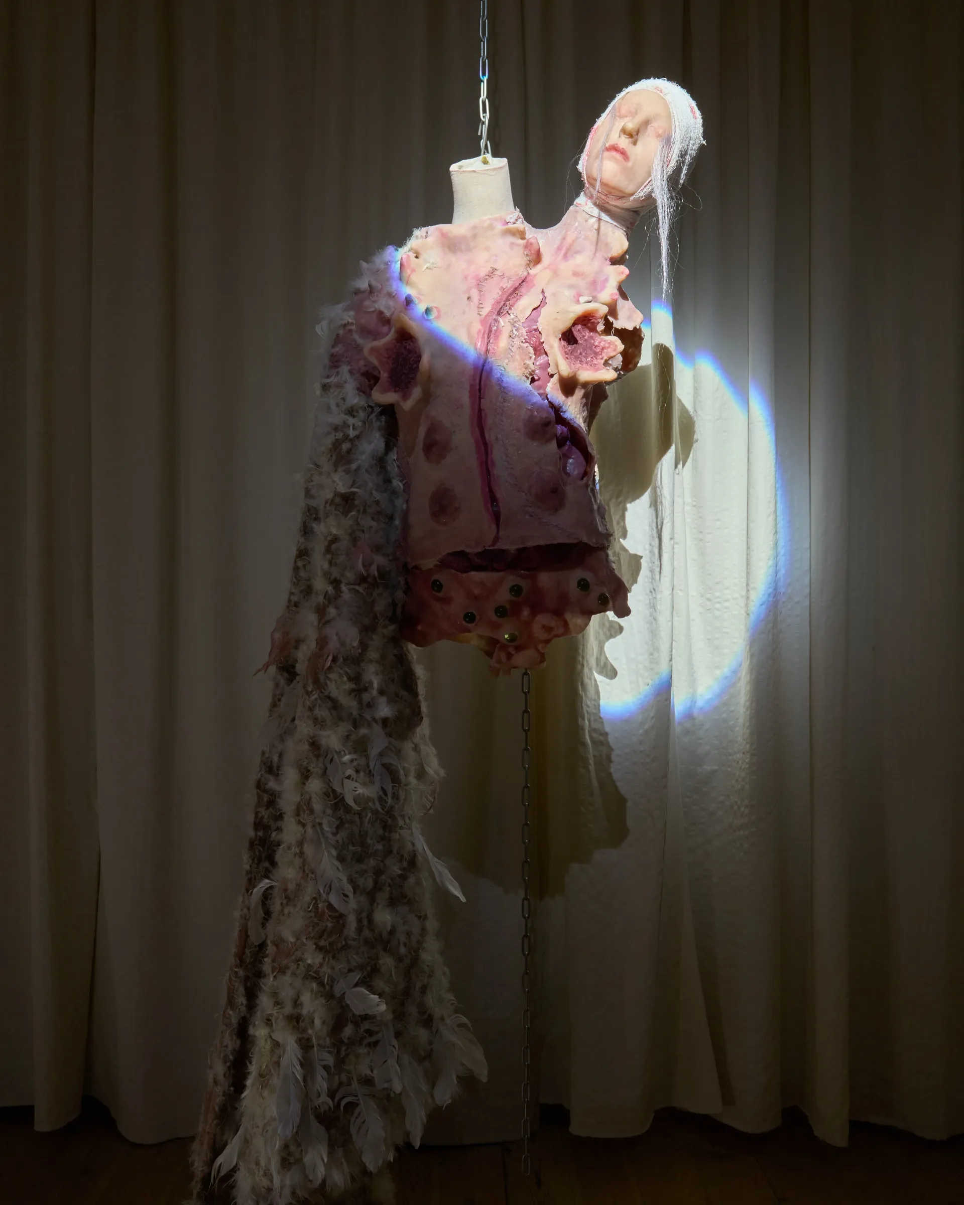Tina Bxtq, Postangelic, Bleed Through, Galerie 35M2, Prague, 2026, hanging costume with sculptural head on mannequin torso against curtain with projected light