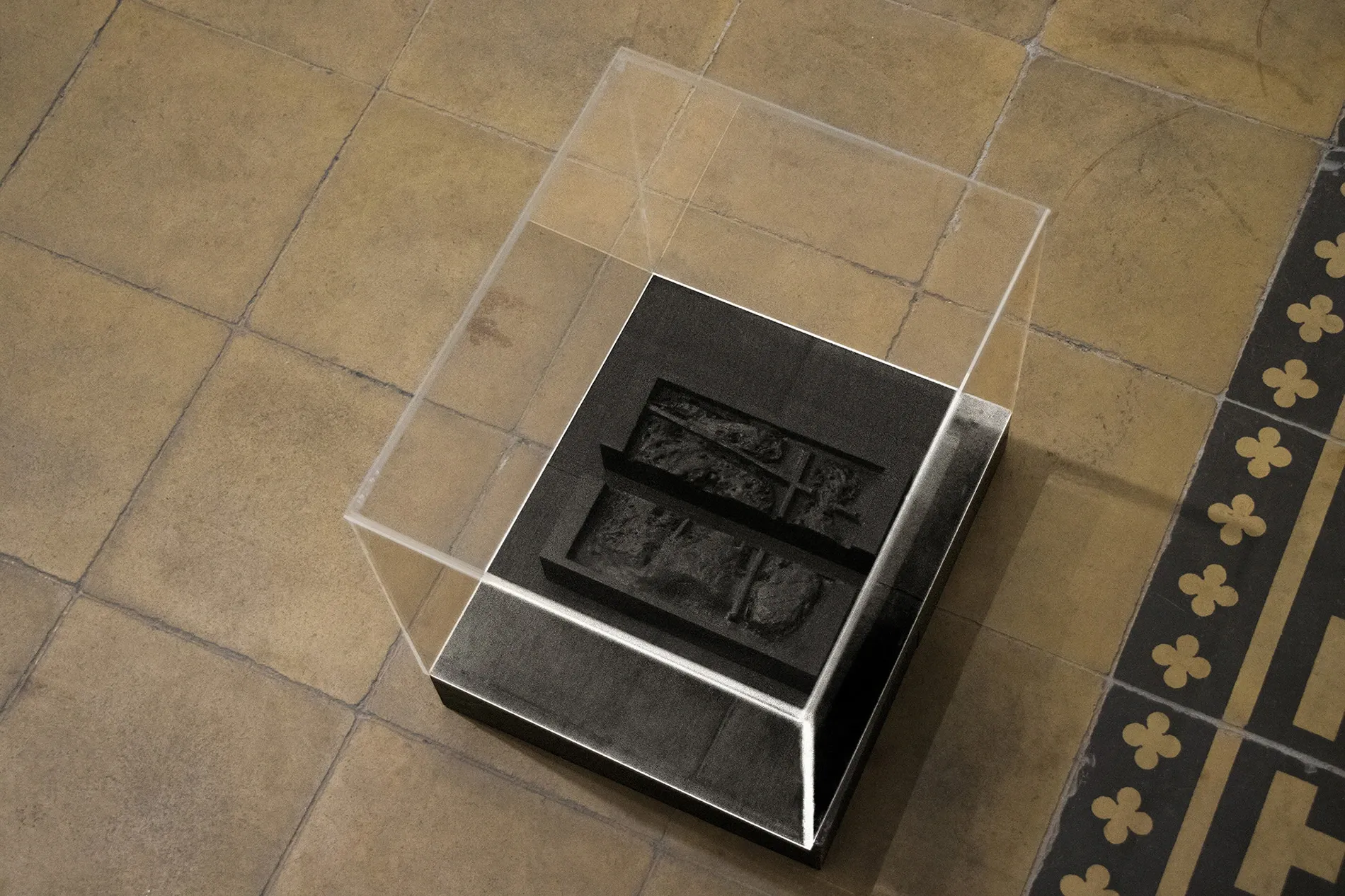 Sofiia Yesakova, Cargo 200, Echoes that refuse to fade, FAVU Gallery Brno, 2026, black sculptural object under plexiglass vitrine on tiled floor