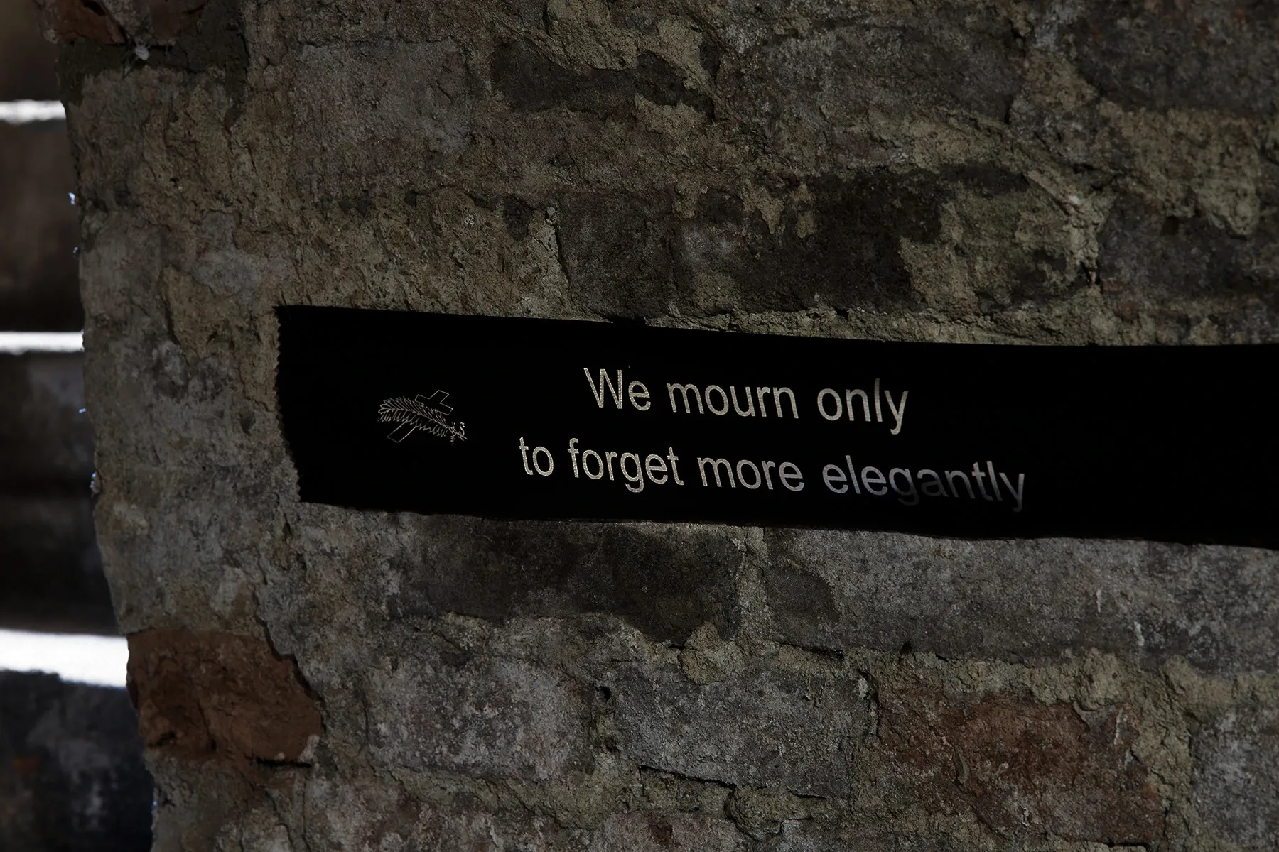 Sofiia Yesakova, Echoes that refuse to fade, FAVU Gallery Brno, 2026, black funeral ribbon reading We mourn only to forget more elegantly on raw brick wall