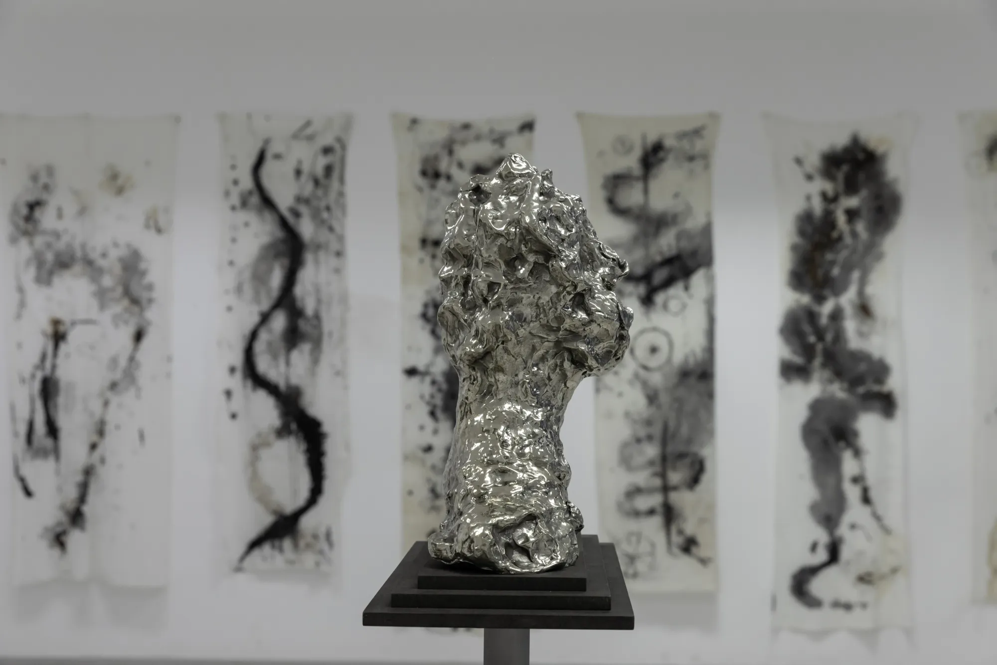 Reflective metal head sculpture on a black pedestal in front of tall black-and-white abstract paintings.