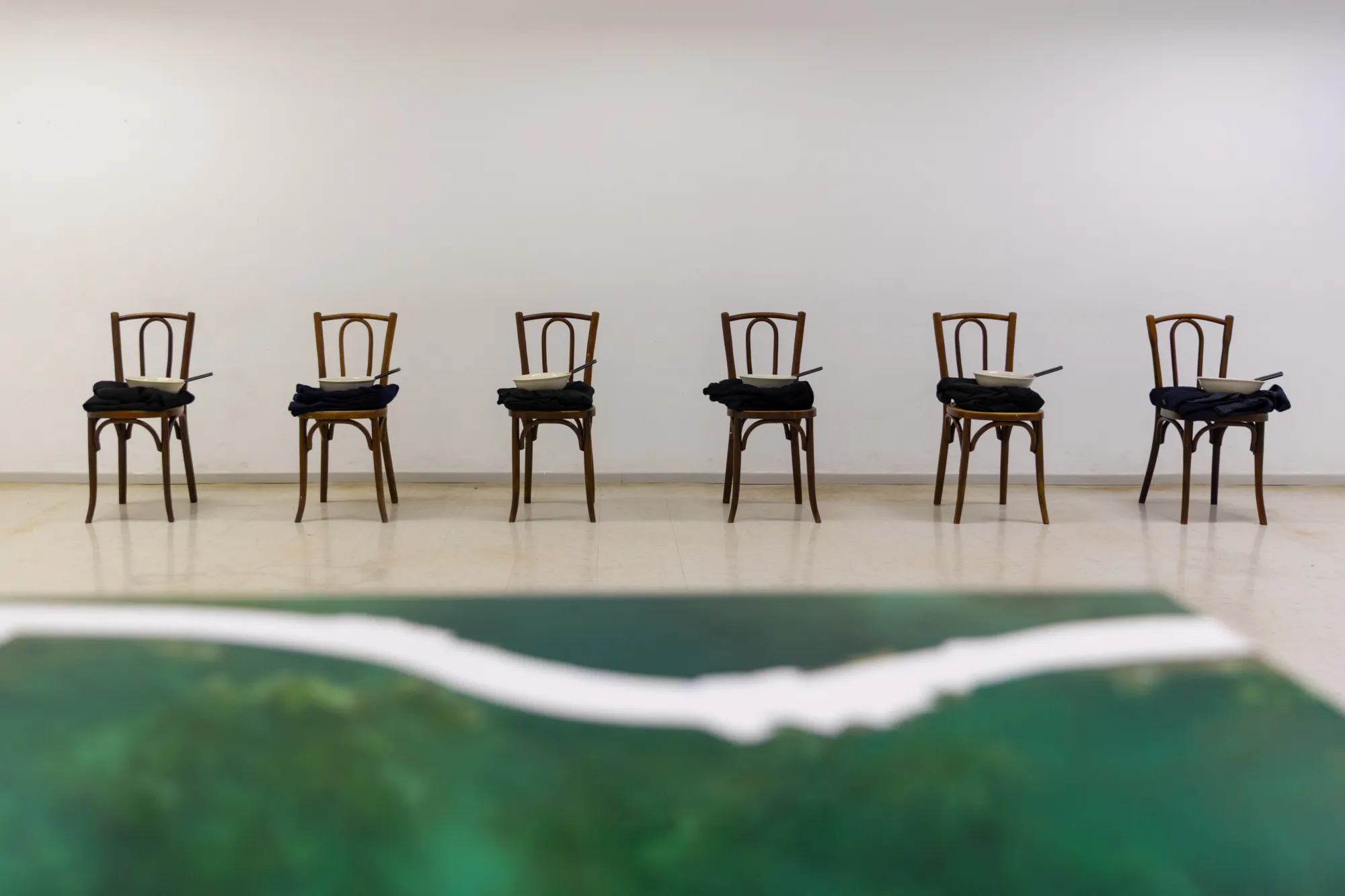 Green resin artwork in the foreground with six wooden chairs holding bowls of water and knives along the gallery wall behind.
