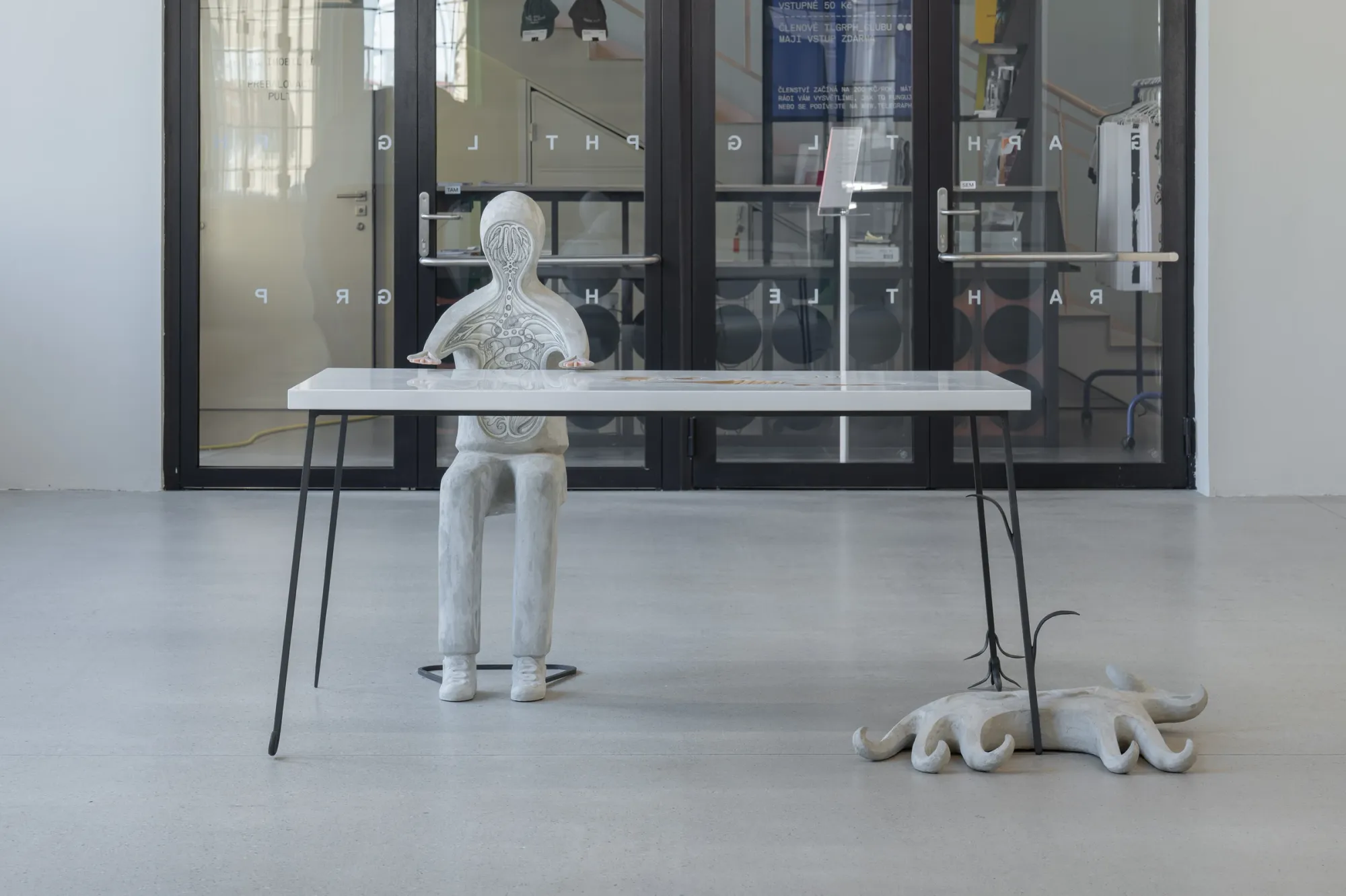 Exhibition review of Anna Hulačová: Signal V at Telegraph Gallery, Olomouc, 2026. Plaster figure seated at a table near the entrance doors.