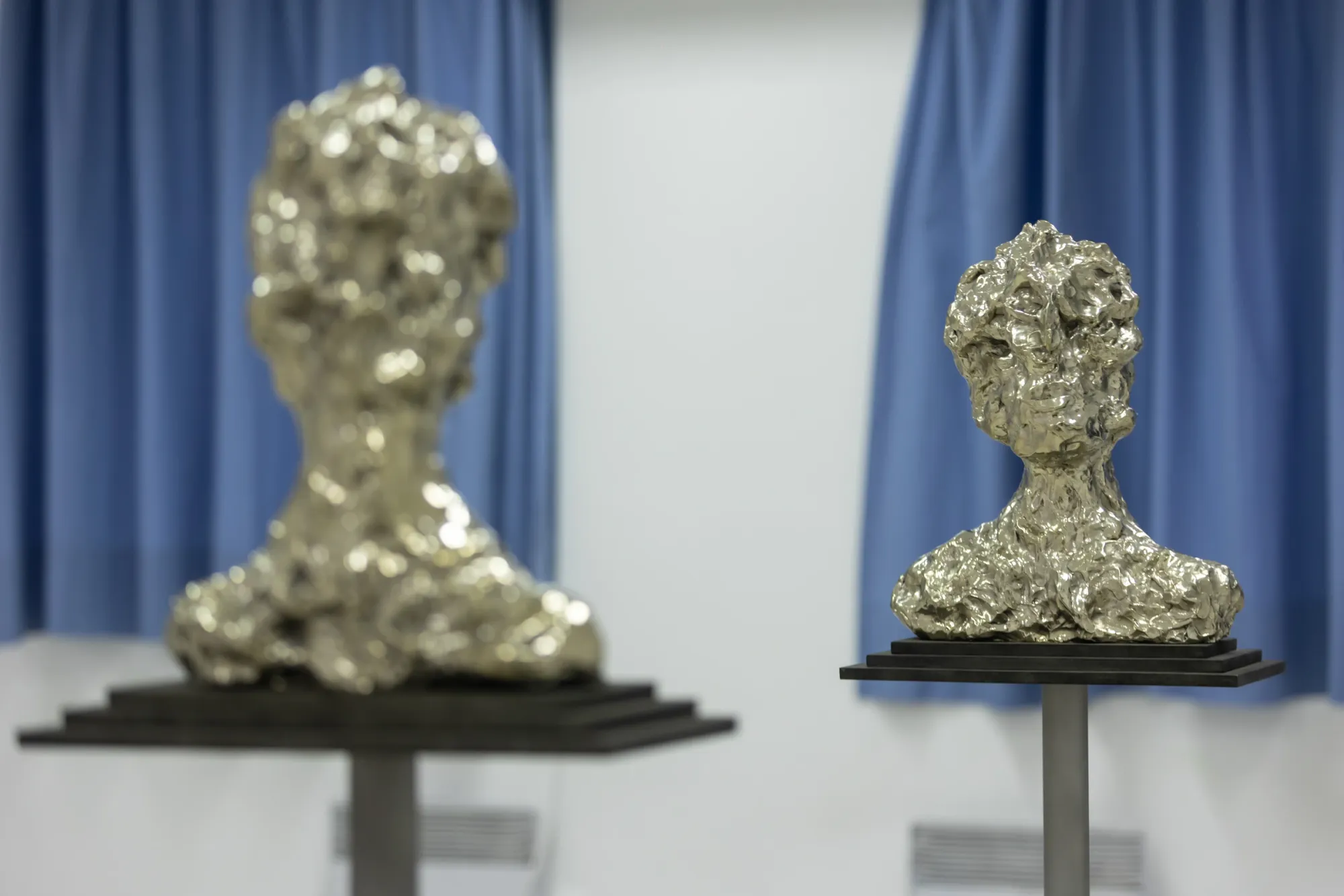 Exhibition review of Daniel Silver, two crumpled reflective metal head sculptures on plinths against a blue curtain, Hydra, 2026.