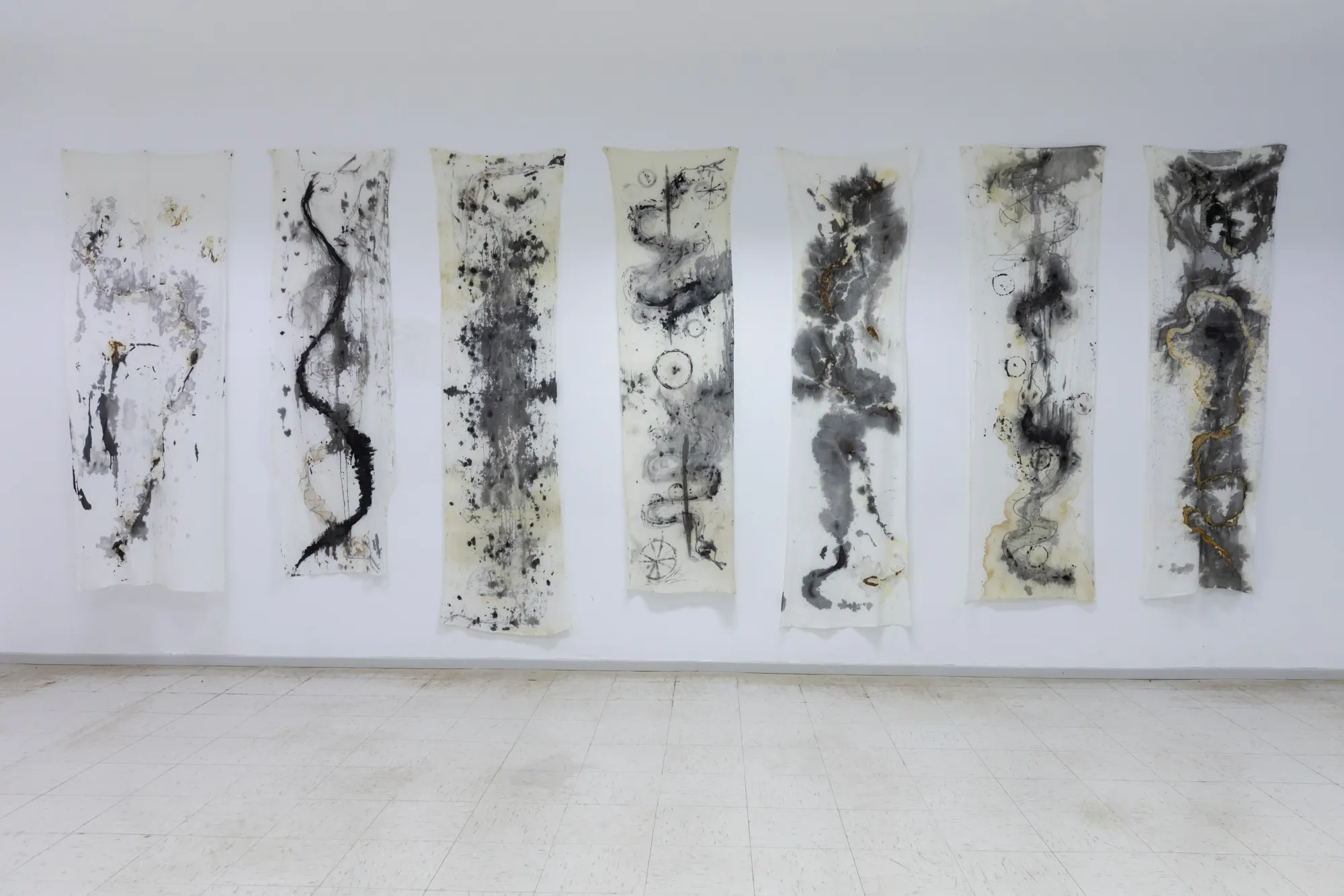 Seven tall abstract works from Dimitrios Antonitsis’s Shrouds series, marked with black, grey and muted gold gestures, installed in a row on a white gallery wall.