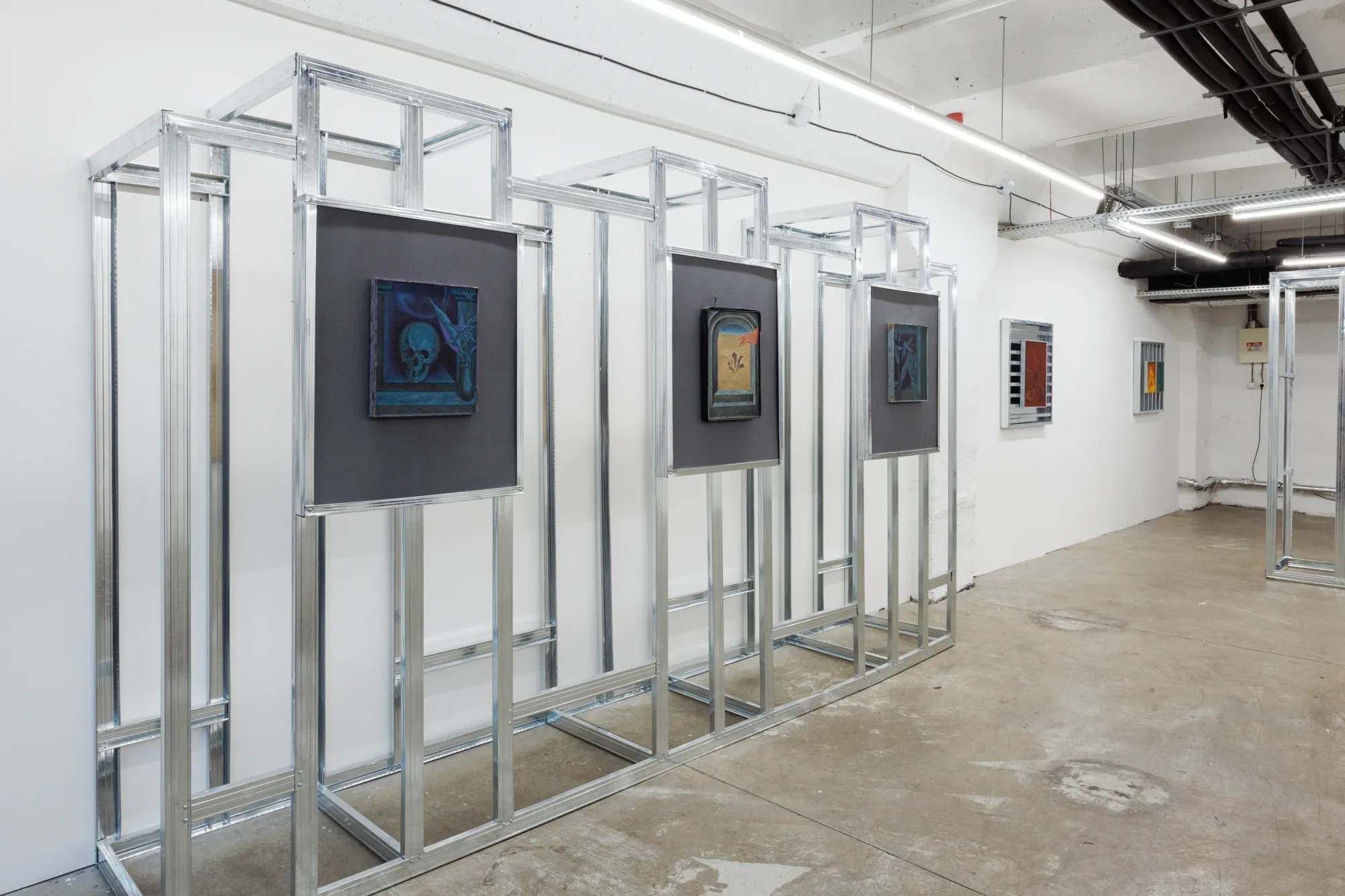 Exhibition review of Erekle Chinchilakashvili: The Story of a Thread Seen From the Window at The Why Not Gallery, Tbilisi, 2026, installation view, diagonal view of the gallery space showing the steel grid structure and smaller framed paintings on the adjacent wall.