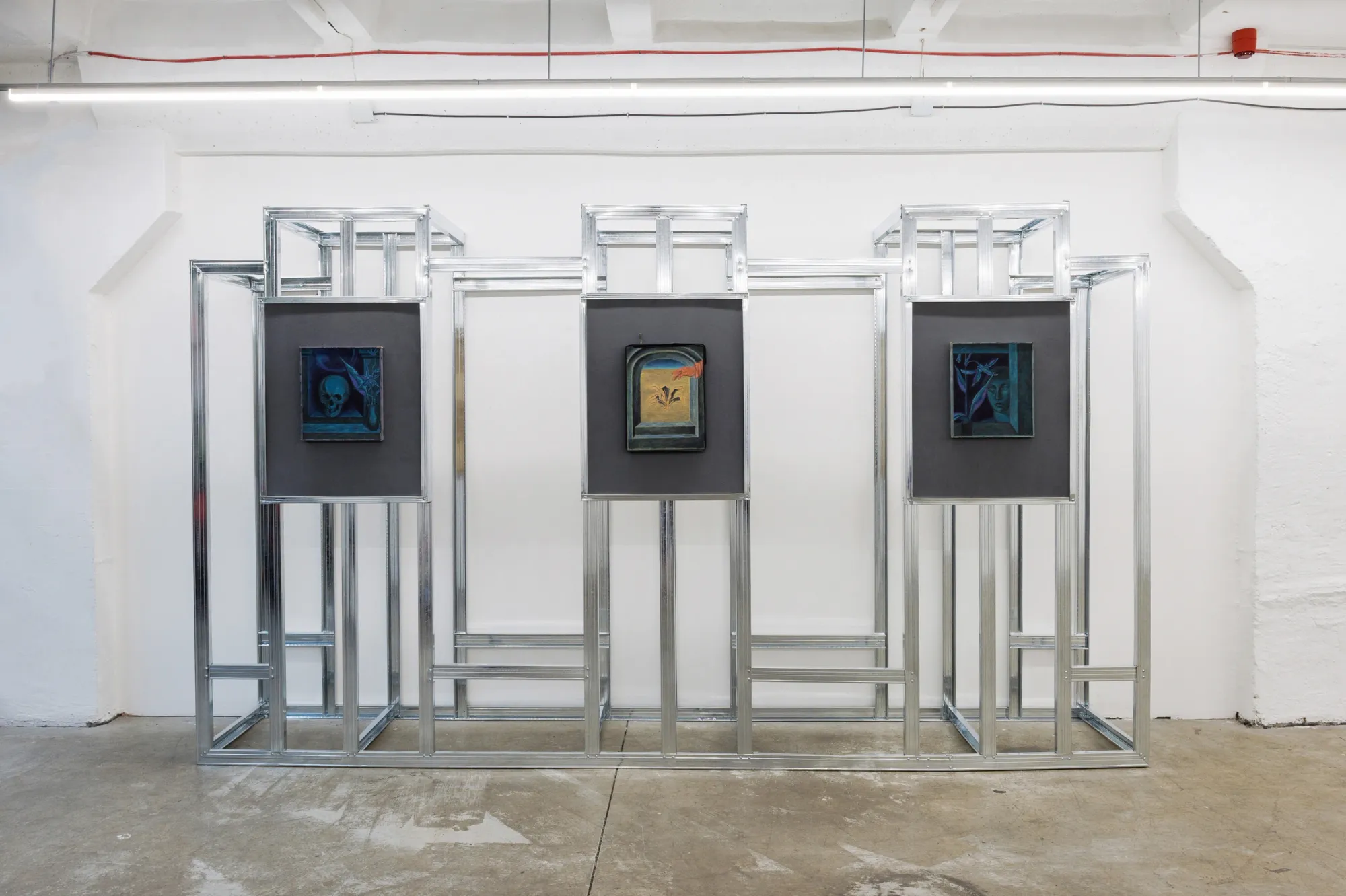 Exhibition review of Erekle Chinchilakashvili: The Story of a Thread Seen From the Window at The Why Not Gallery, Tbilisi, 2026, installation view, three small paintings in dark frames mounted in a row within an aluminum grid structure.