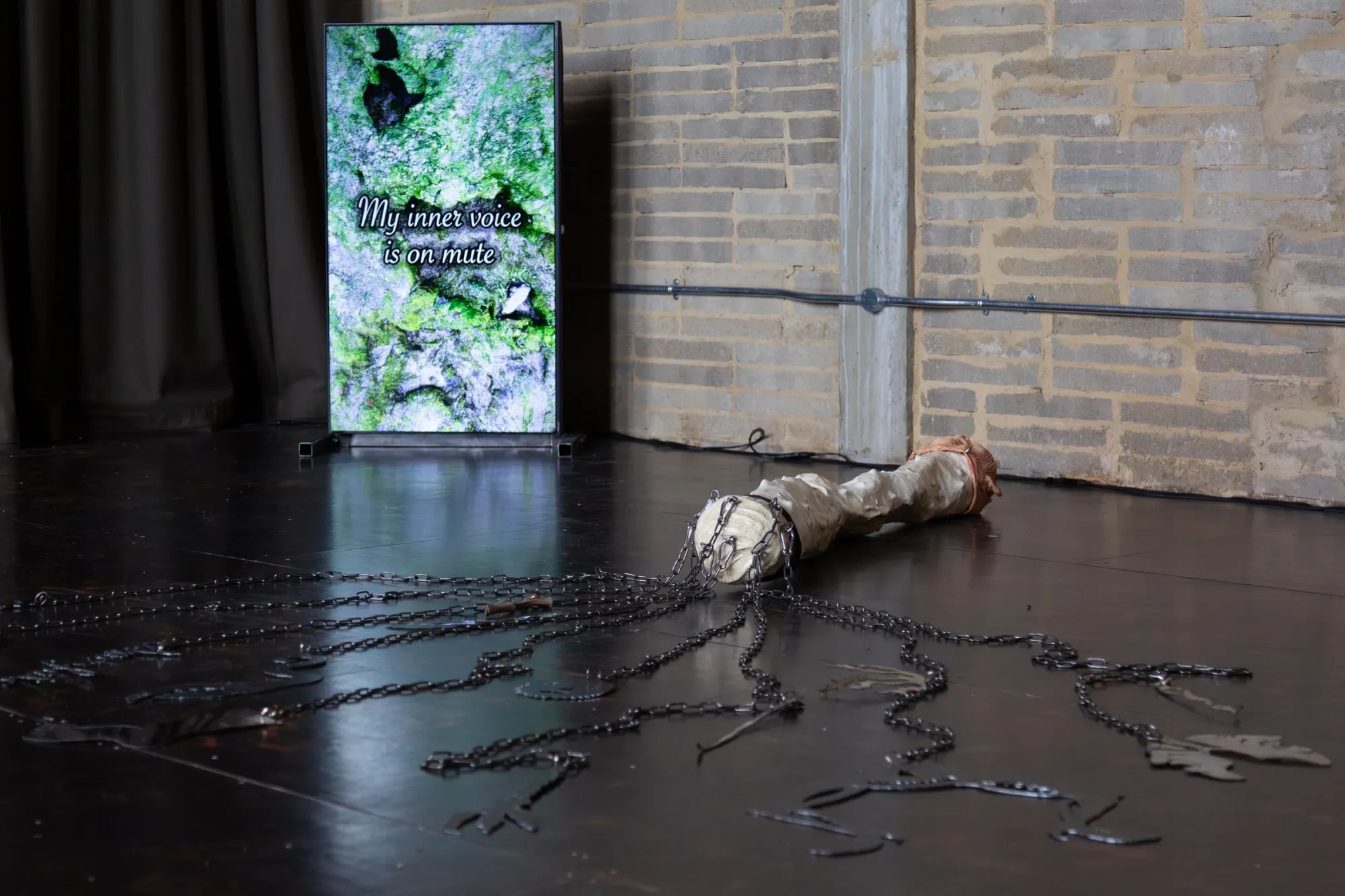 Nicola Genovese, Infinite Stage, detail, cast limb sculpture in chains before a screen reading My inner voice is on mute, Raleigh Chapel, 2026