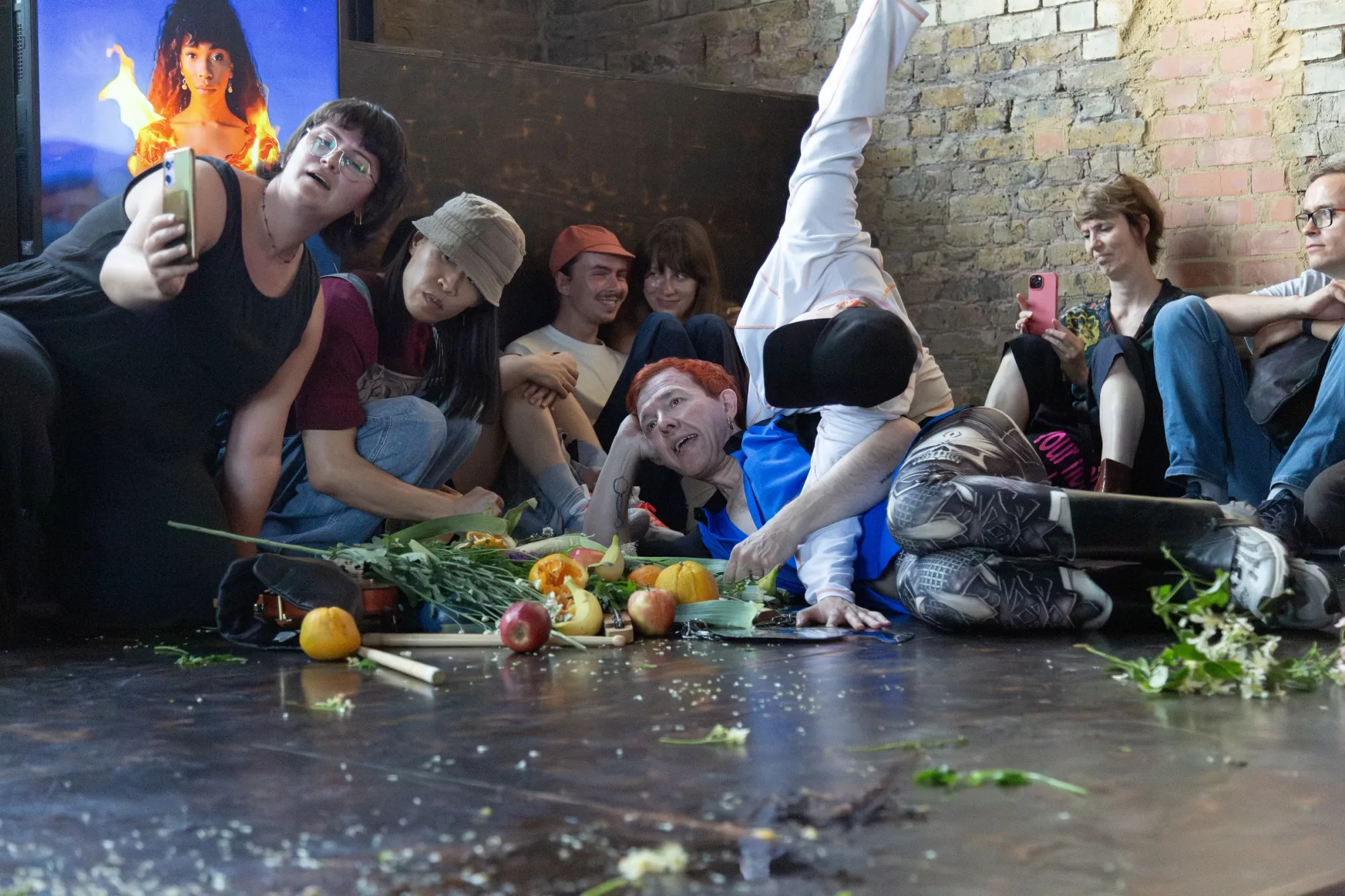 Nicola Genovese, Infinite Stage, Raleigh Chapel, London, 2026, audience recording on phones as a performer lies among scattered fruit and flowers