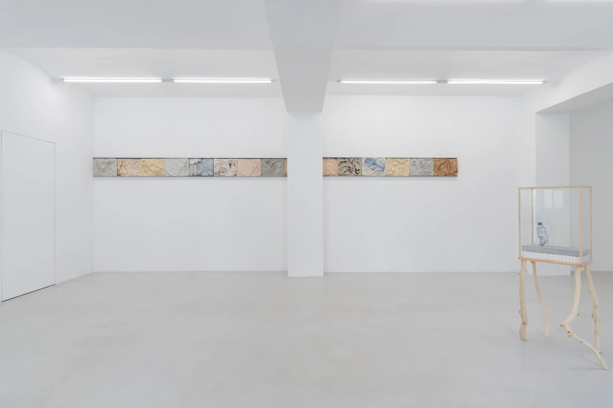 Exhibition review of Agnieszka Topolska: Call It Fate, Call It Karma at Galeria Skala, Poznań, 2026. Row of six textured cast relief tiles in peach, grey, blue, cream and sand tones.