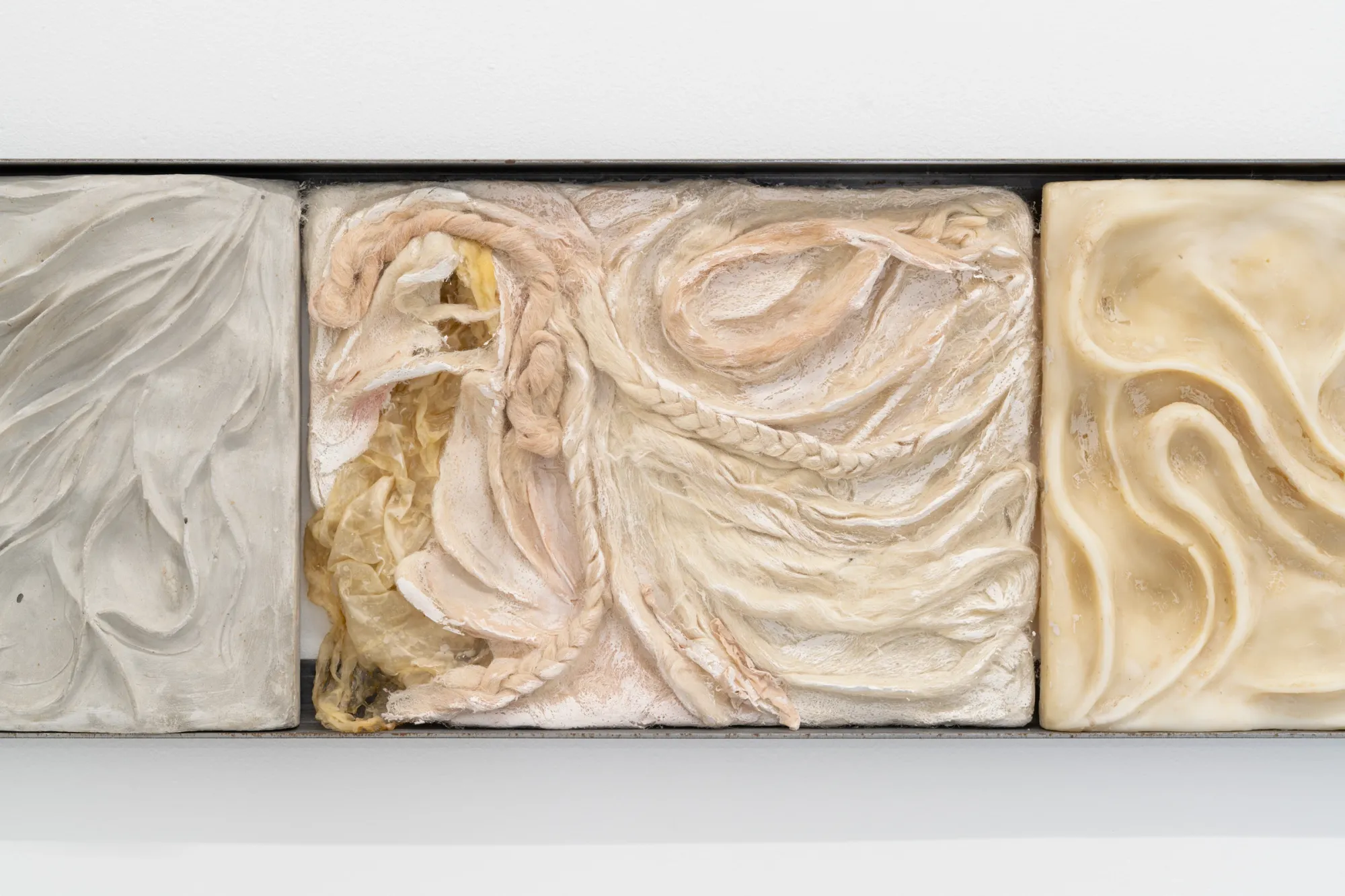 Exhibition review of Agnieszka Topolska: Call It Fate, Call It Karma at Galeria Skala, Poznań, 2026. Close-up of two cast relief tiles, one carved plaster fold pattern, one with pale fabric embedded in wax.