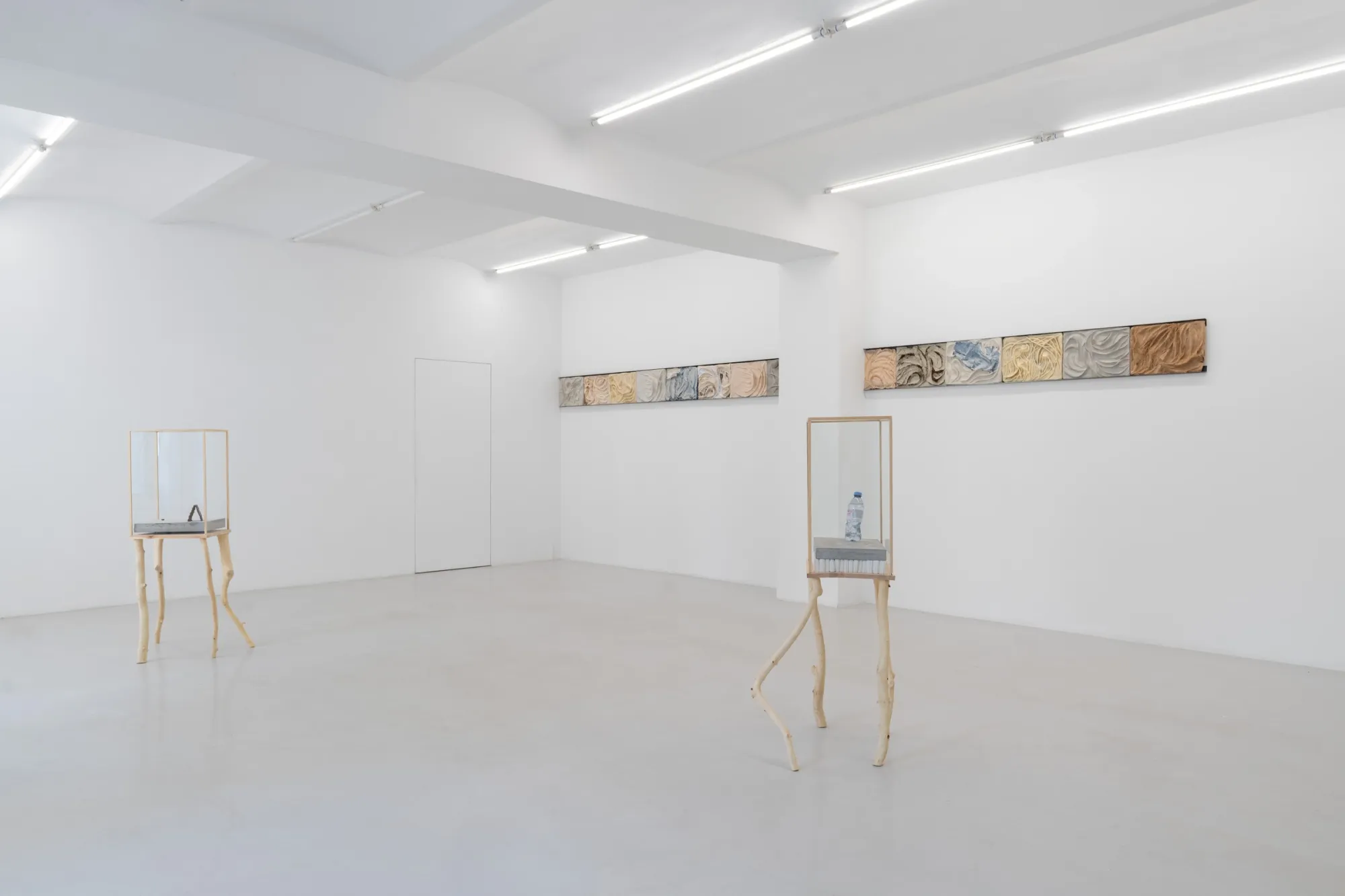 Exhibition review of Agnieszka Topolska: Call It Fate, Call It Karma at Galeria Skala, Poznań, 2026. Corner gallery view with two long textured wall relief rows and two glass display cases.