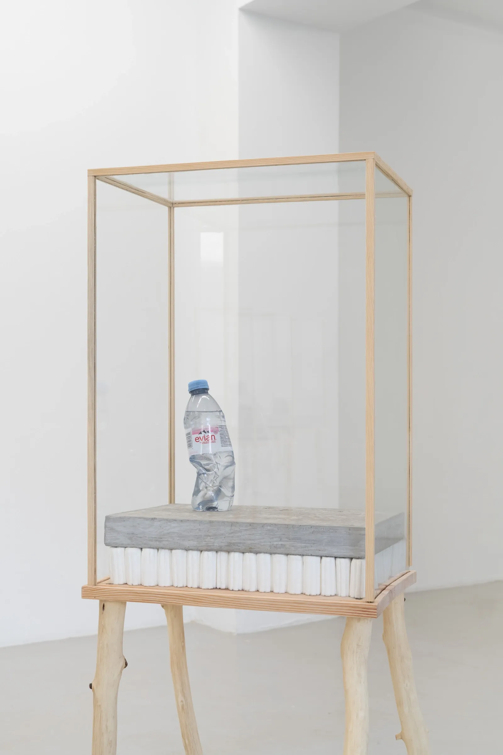 Exhibition review of Agnieszka Topolska: Call It Fate, Call It Karma at Galeria Skala, Poznań, 2026. Close view of a crushed clear plastic Evian water bottle standing on a concrete slab inside a glass case.