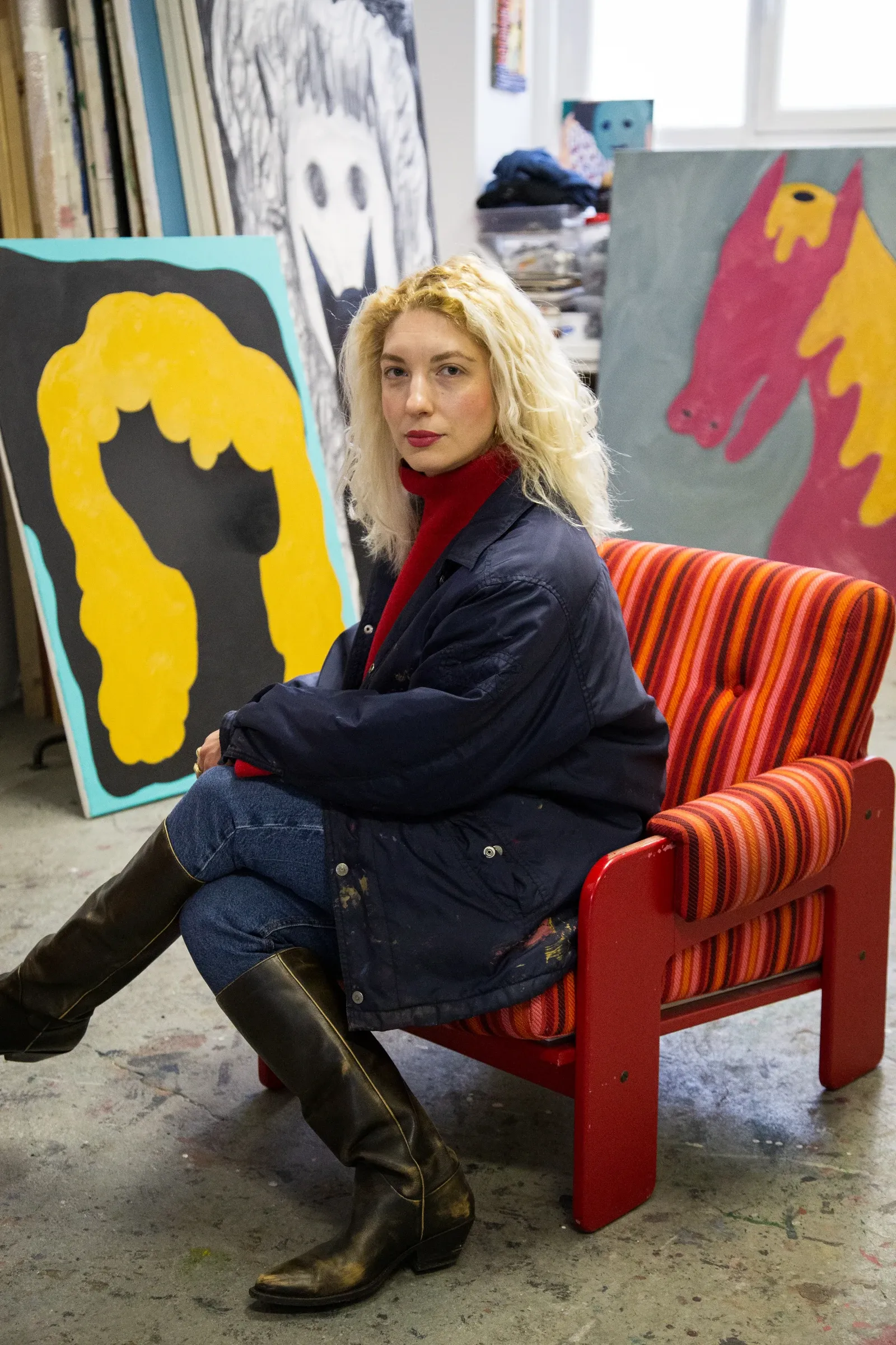 Exhibition review of Kallirroi Ioannidou: Galvanizing Memories at Kallmann-Museum, Ismaning, 2026. The artist seated in her studio among paintings, including a yellow wig-shaped canvas and a pink and yellow horse head.
