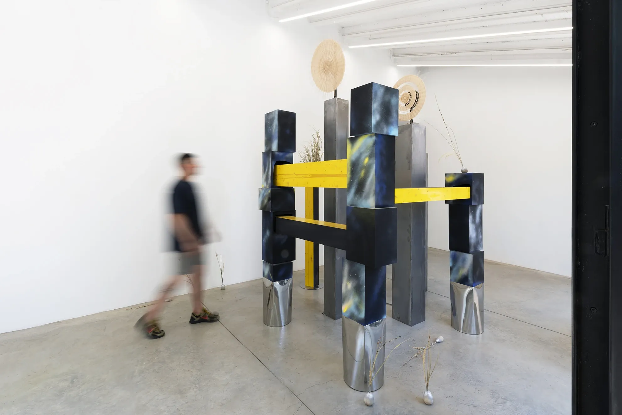 Exhibition review of Leah Dixon: Tartan Tartana at Spiritvessel, Espinavessa, 2026. A visitor walks past the steel and yellow wood sculpture in a sunlit gallery.