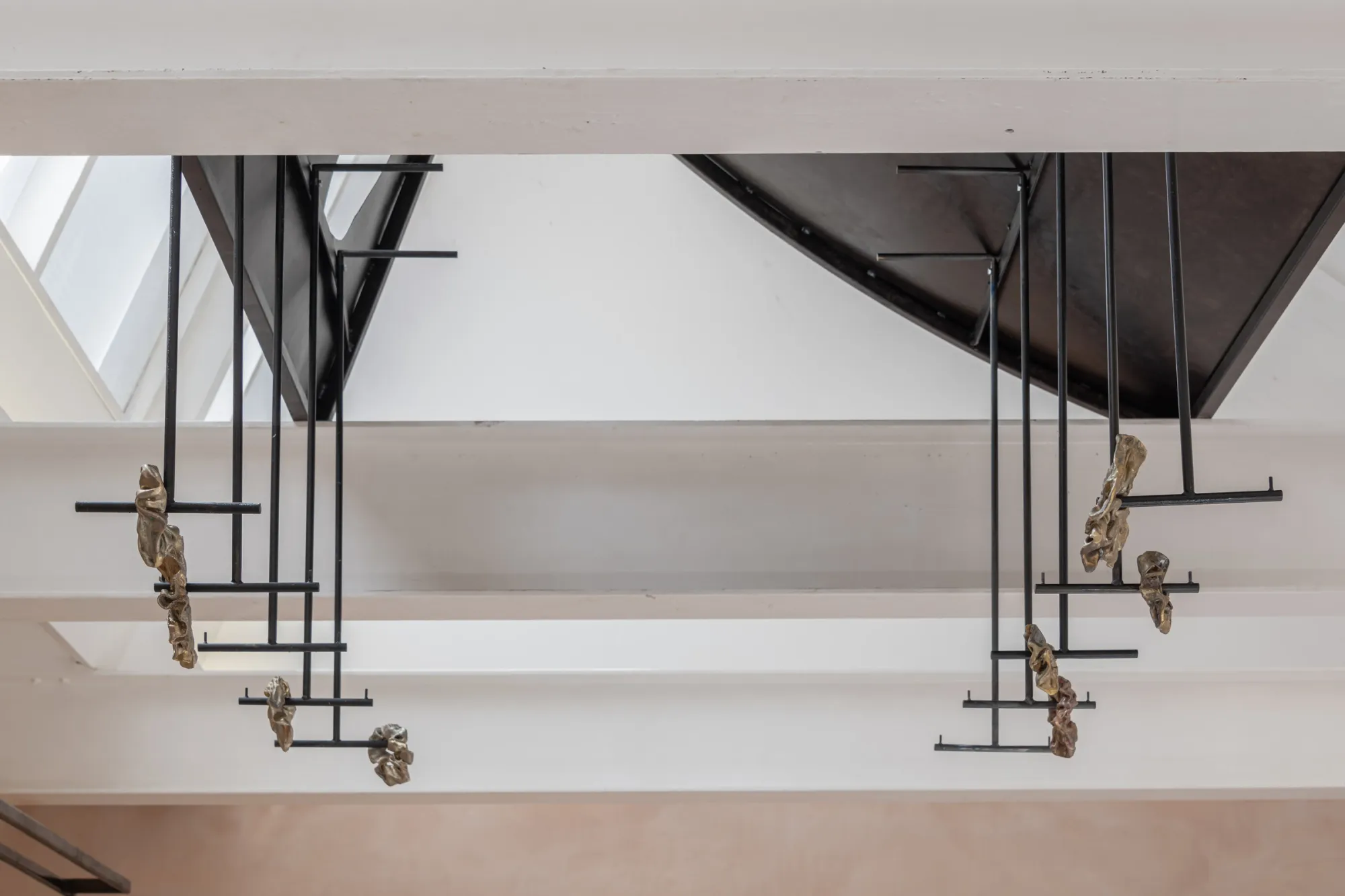 Exhibition review of Maija Fox: As the sap rises at OUTPOST, Norwich, 2026. Cast brass forms hanging on wire brackets beneath a black triangular roof structure.