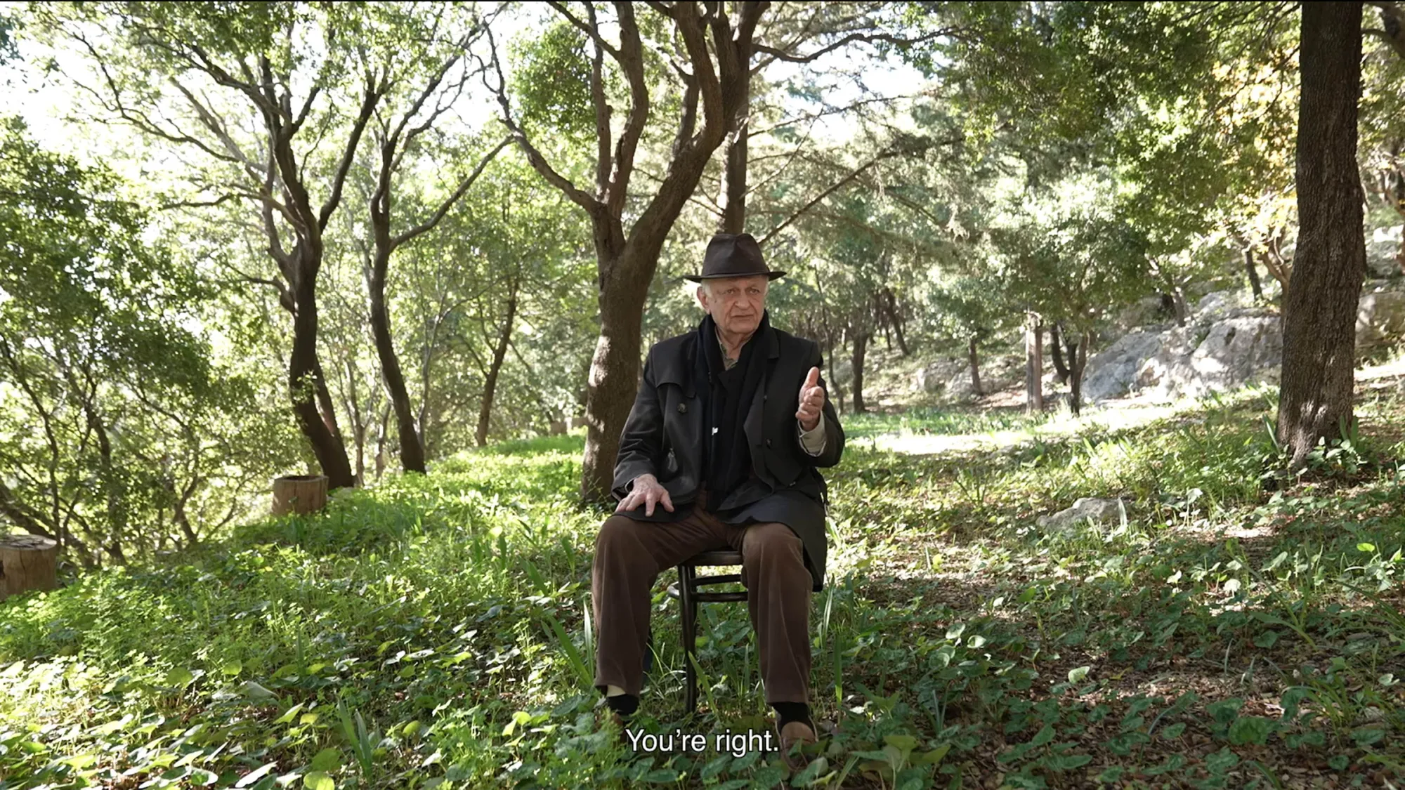 Exhibition review of Marwa Arsanios: Who is Afraid of Ideology? at TAXISPALAIS Kunsthalle Tirol, Innsbruck, 2026. Elderly man in a hat seated on a chair among trees and rocks in a wooded landscape.