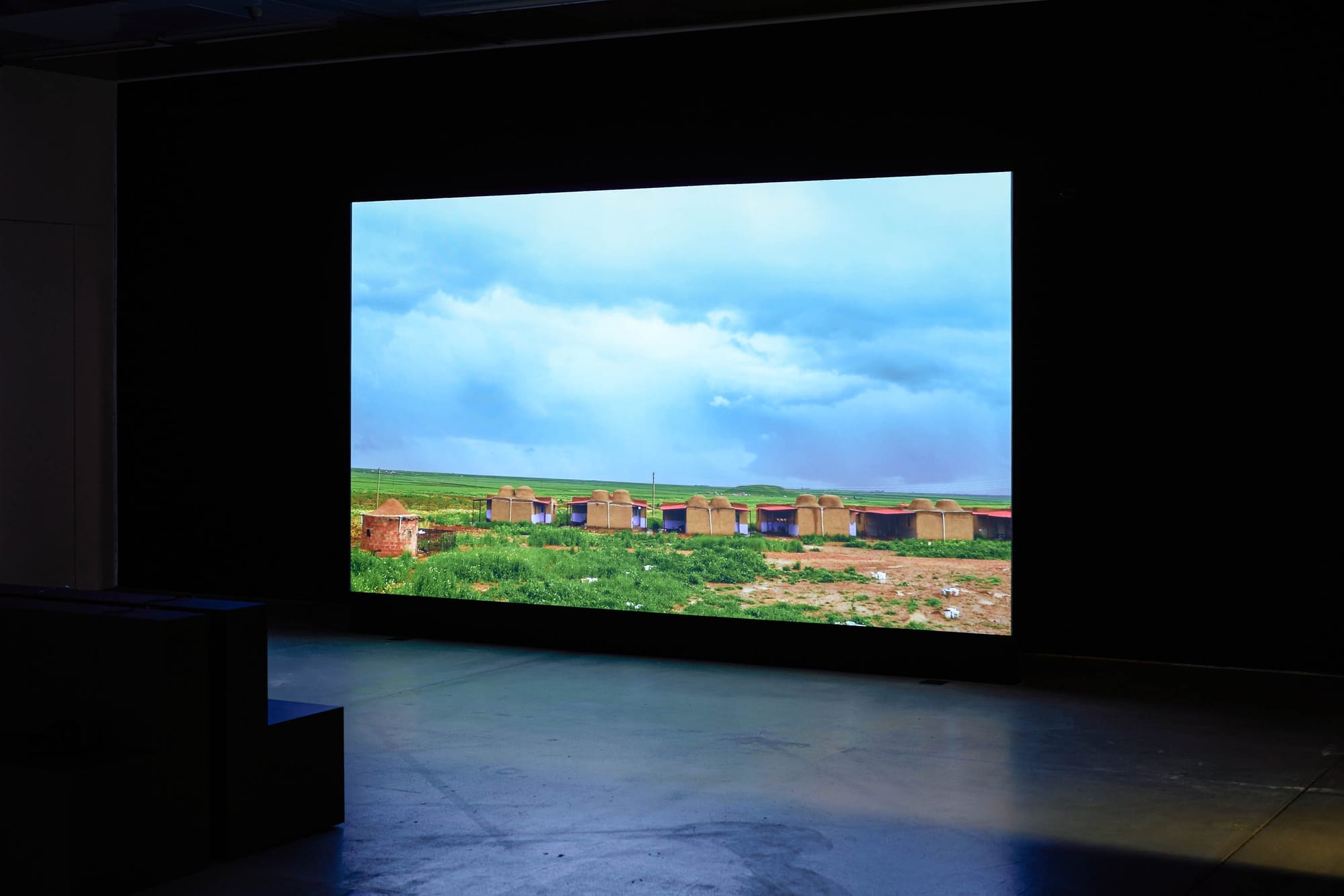 Exhibition review of Marwa Arsanios: Who is Afraid of Ideology? at TAXISPALAIS Kunsthalle Tirol, Innsbruck, 2026. Screen showing mud brick huts with red roofs in a green field under a stormy sky.