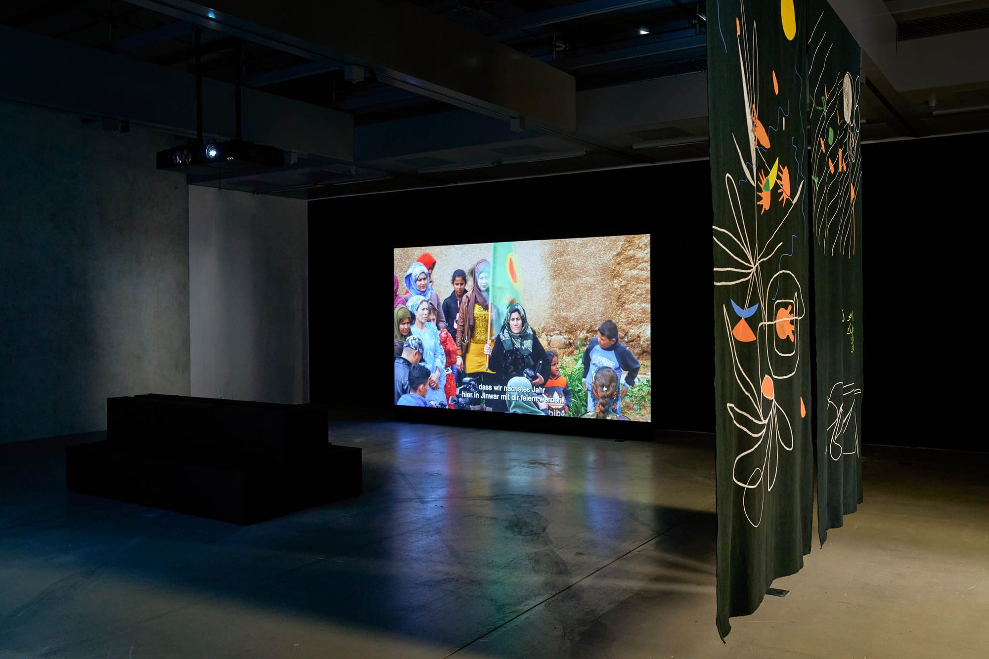 catapult contemporary Exhibition review of Marwa Arsanios: Who is Afraid of Ideology? at TAXISPALAIS Kunsthalle Tirol, Innsbruck, 2026. Wide screen showing a crowd holding a yellow flag beside a dark printed textile drape.