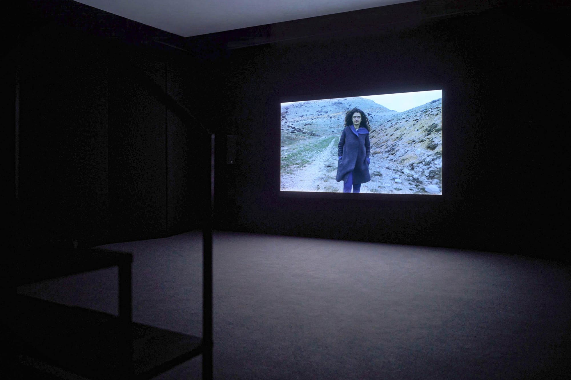 Exhibition review of Marwa Arsanios: Who is Afraid of Ideology? at TAXISPALAIS Kunsthalle Tirol, Innsbruck, 2026. Dark room with a single wall screen showing a woman standing on a mountain path.