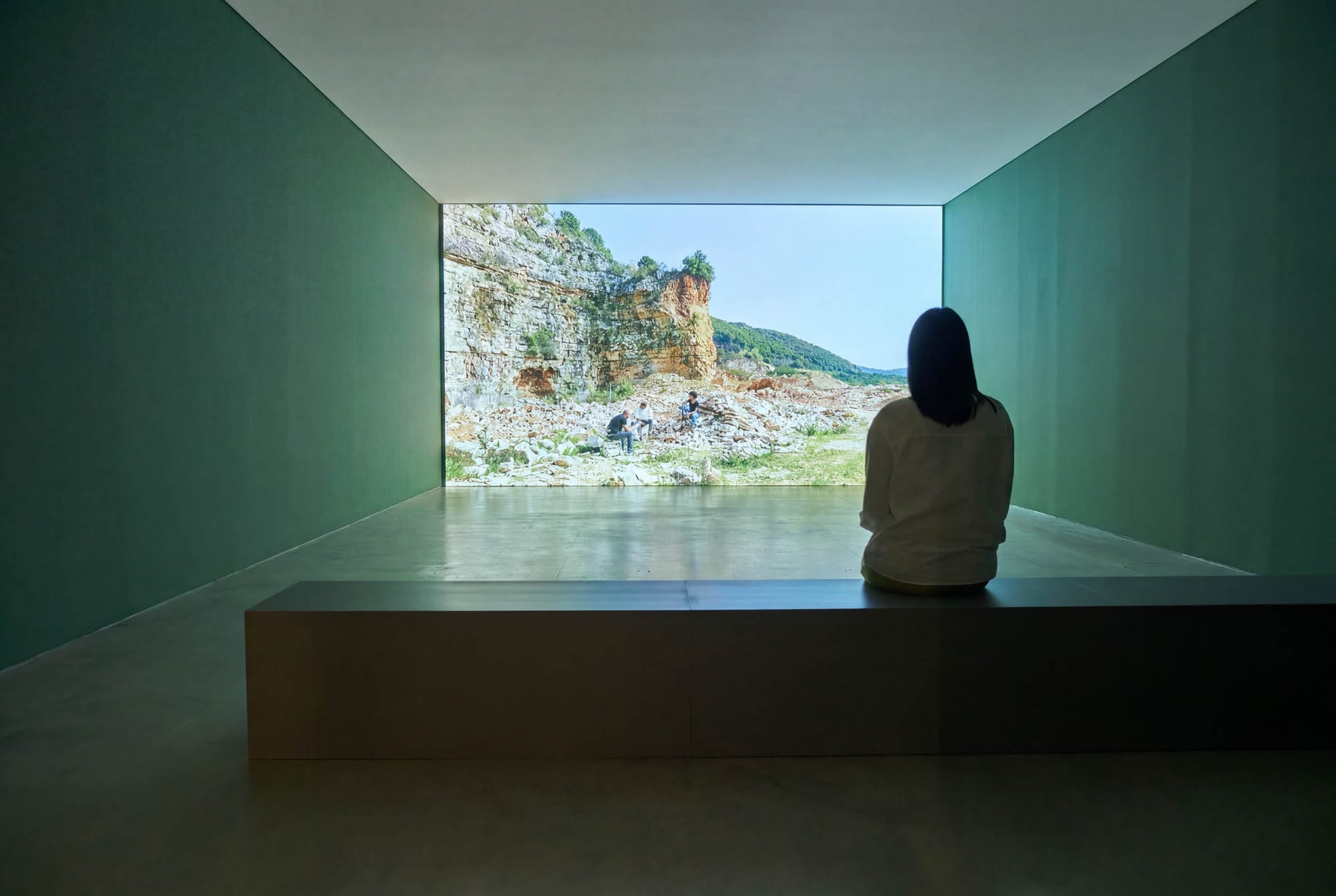 Exhibition review of Marwa Arsanios: Who is Afraid of Ideology? at TAXISPALAIS Kunsthalle Tirol, Innsbruck, 2026. Green room with a low bench facing a screen of a quarry where people sit among rubble.