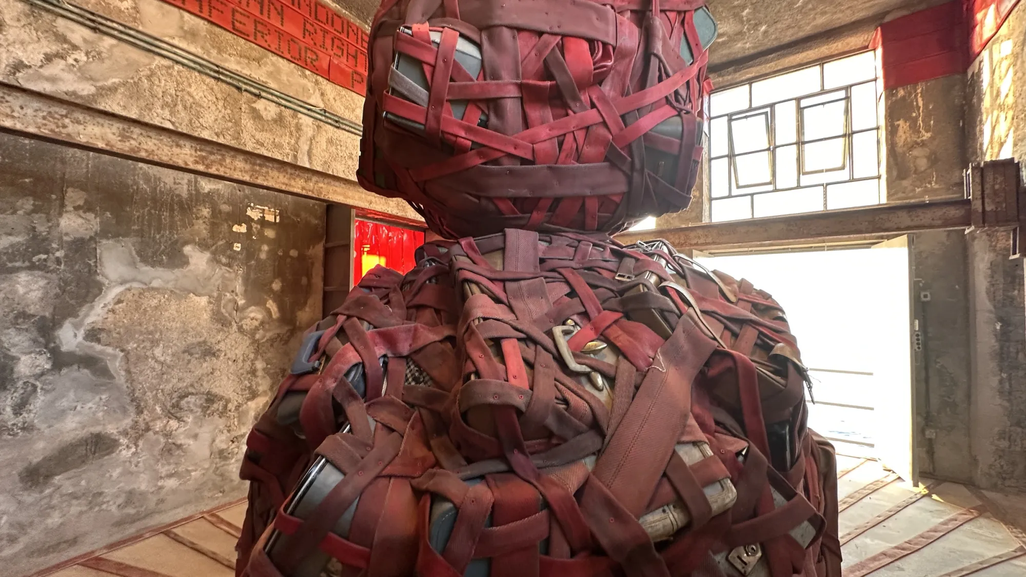 Exhibition review of Nari Ward: Until That Day at DESTE Foundation, Slaughterhouse, Hydra, 2026. Close view of the woven totem's head, dark red straps crossed over a rounded form.