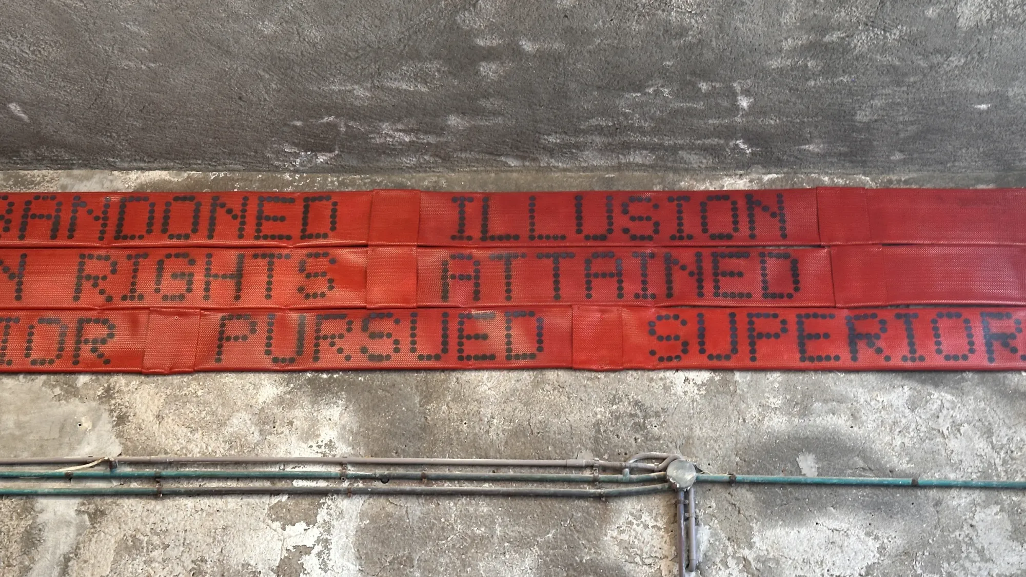 Exhibition review of Nari Ward: Until That Day at DESTE Foundation, Slaughterhouse, Hydra, 2026. Close view of a red banner with raised dot-pattern lettering spelling speech fragments.