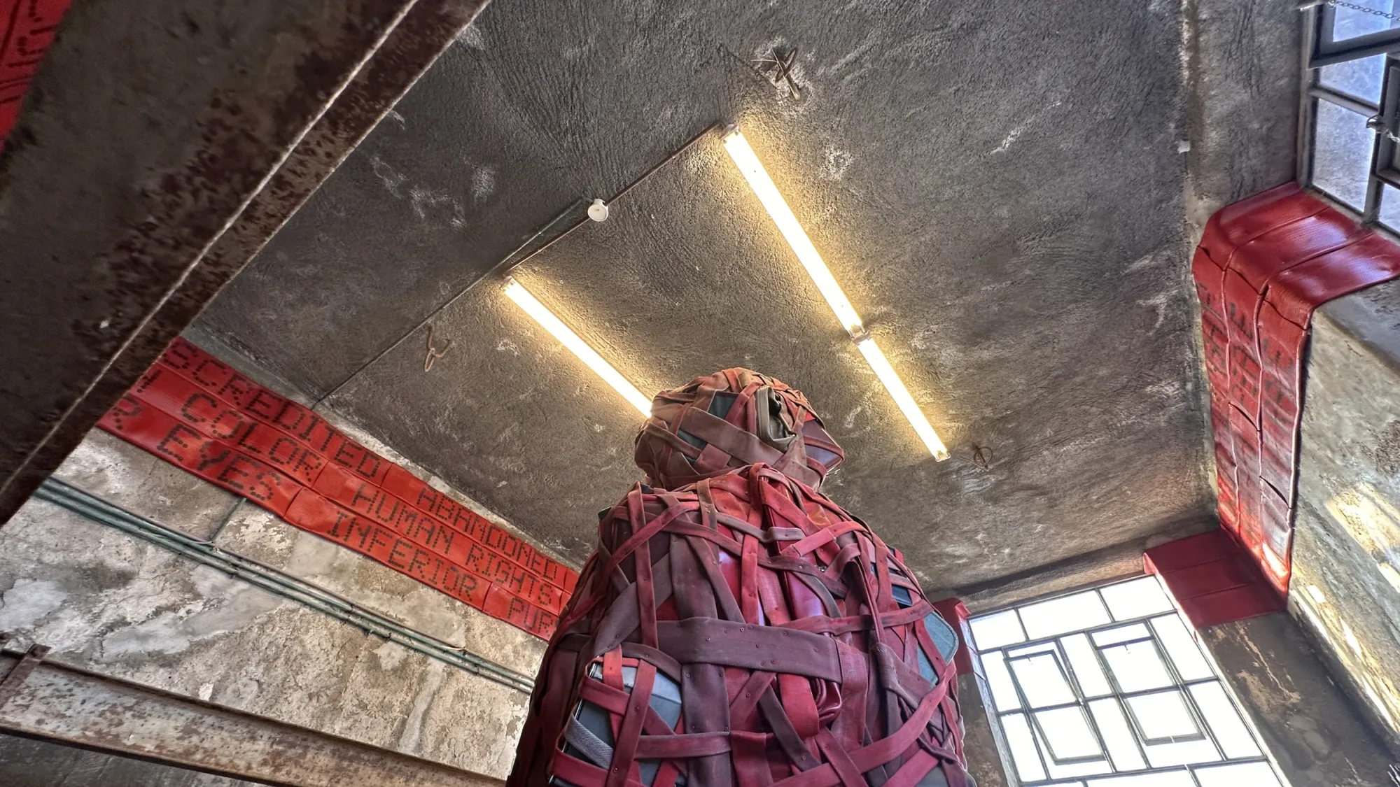 Exhibition review of Nari Ward: Until That Day at DESTE Foundation, Slaughterhouse, Hydra, 2026. A towering red woven strap figure bound around packed luggage under fluorescent light.