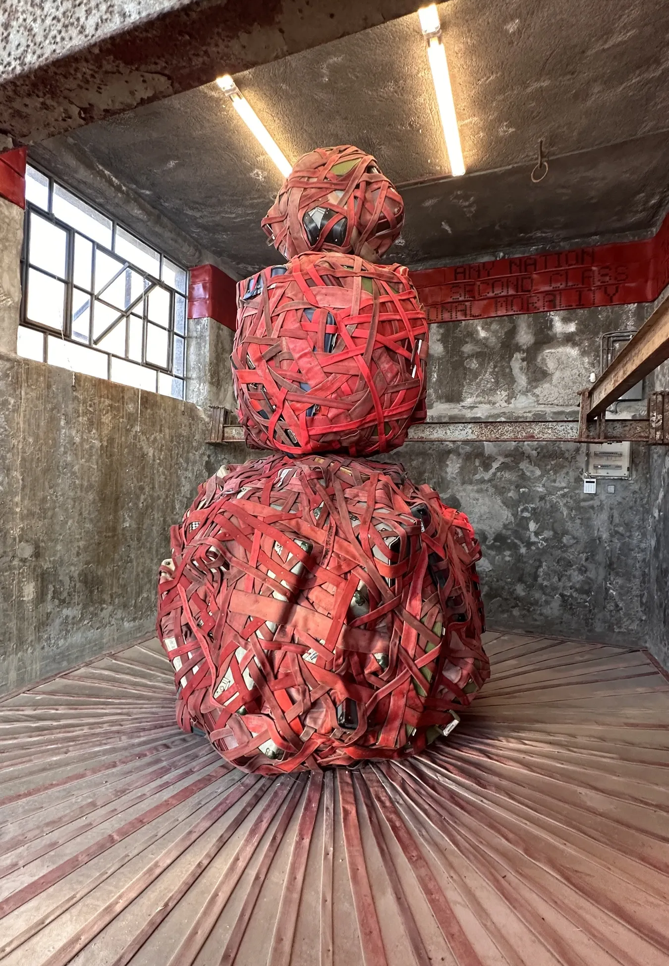 Catapult contemporary art platform Exhibition review of Nari Ward: Until That Day at DESTE Foundation, Slaughterhouse, Hydra, 2026. A red strap-bound totem beneath a raised red text banner quoting a 1963 speech.