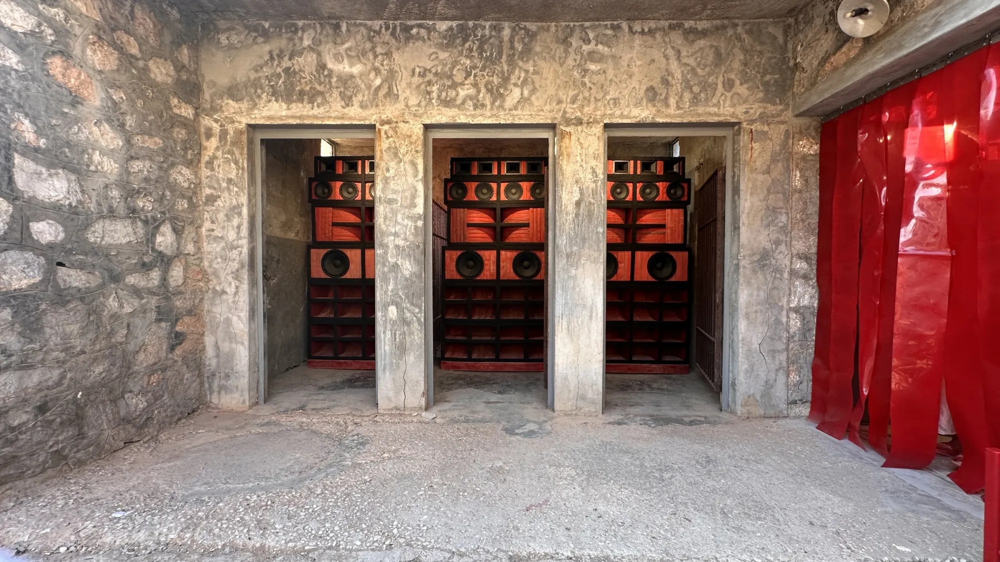 Exhibition review of Nari Ward: Until That Day at DESTE Foundation, Slaughterhouse, Hydra, 2026. Three matching red and black sound system towers seen through stone doorways.