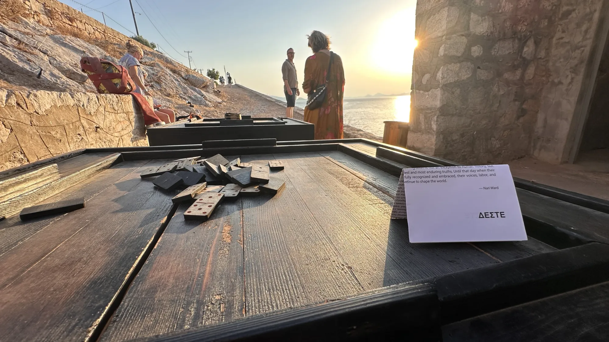 Exhibition review of Nari Ward: Until That Day at DESTE Foundation, Slaughterhouse, Hydra, 2026. A dark wood domino table holding a printed quote card by the artist, sea behind.