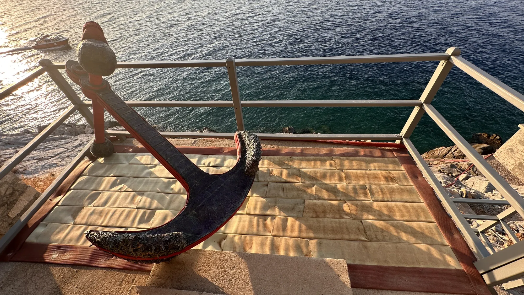 Exhibition review of Nari Ward: Until That Day at DESTE Foundation, Slaughterhouse, Hydra, 2026. A ship's anchor wrapped in red woven strapping on a terrace above the sea.