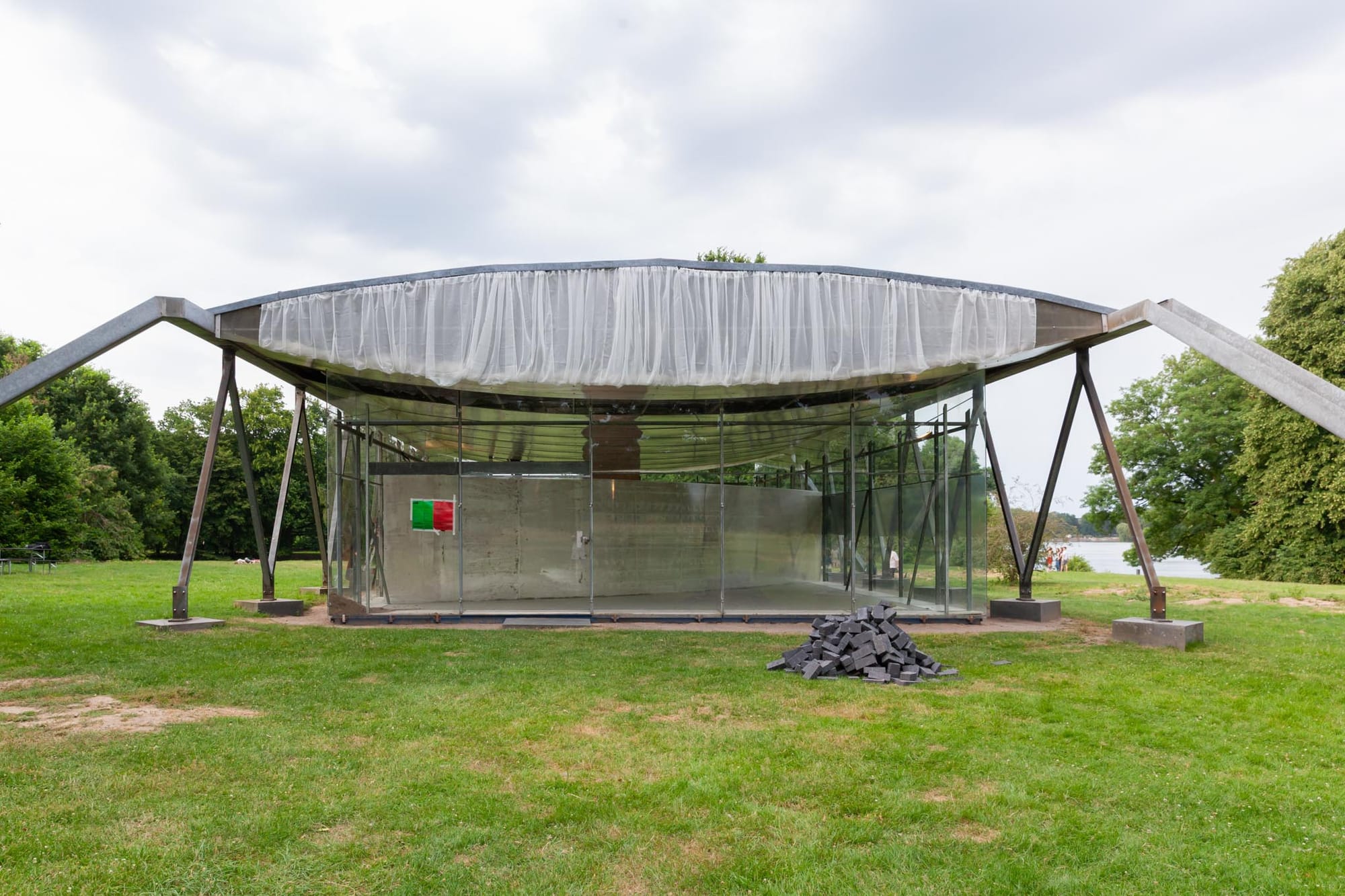 Reparaturgesellschaft, Wewerka Pavilion, Münster, 2026, white curtain draped along pavilion roof edge with pile of dark stone pavers on grass