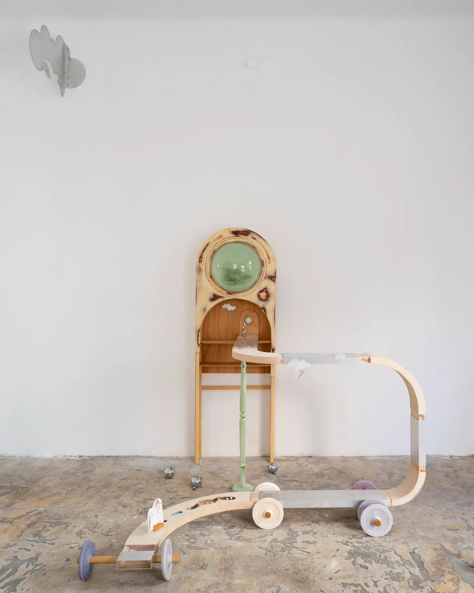 Exhibition review of Sebastian Scholz: ludo at global international, Vienna, 2026. Corner view with a drawing-covered waggon, a green glass porthole cabinet and a wheeled floor sculpture.