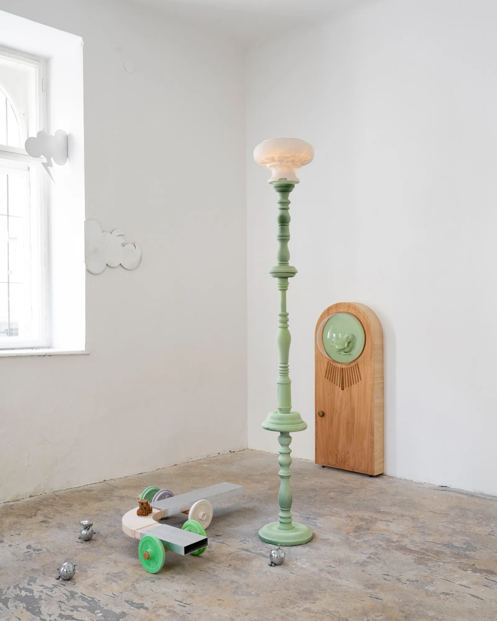 Exhibition review of Sebastian Scholz: ludo at global international, Vienna, 2026. Vertical view of a steel cloud sconce above a wooden cabinet with a green glass porthole.