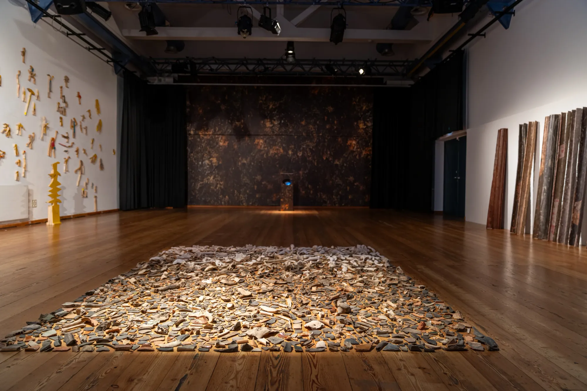 catapult contemporary Exhibition review of TRANSFER at Kulturforum Laßnitzhaus, Deutschlandsberg, 2026. Frontal room view with a wall of wood cutout figures, a floor installation and leaning cast pipes under a rigged ceiling.