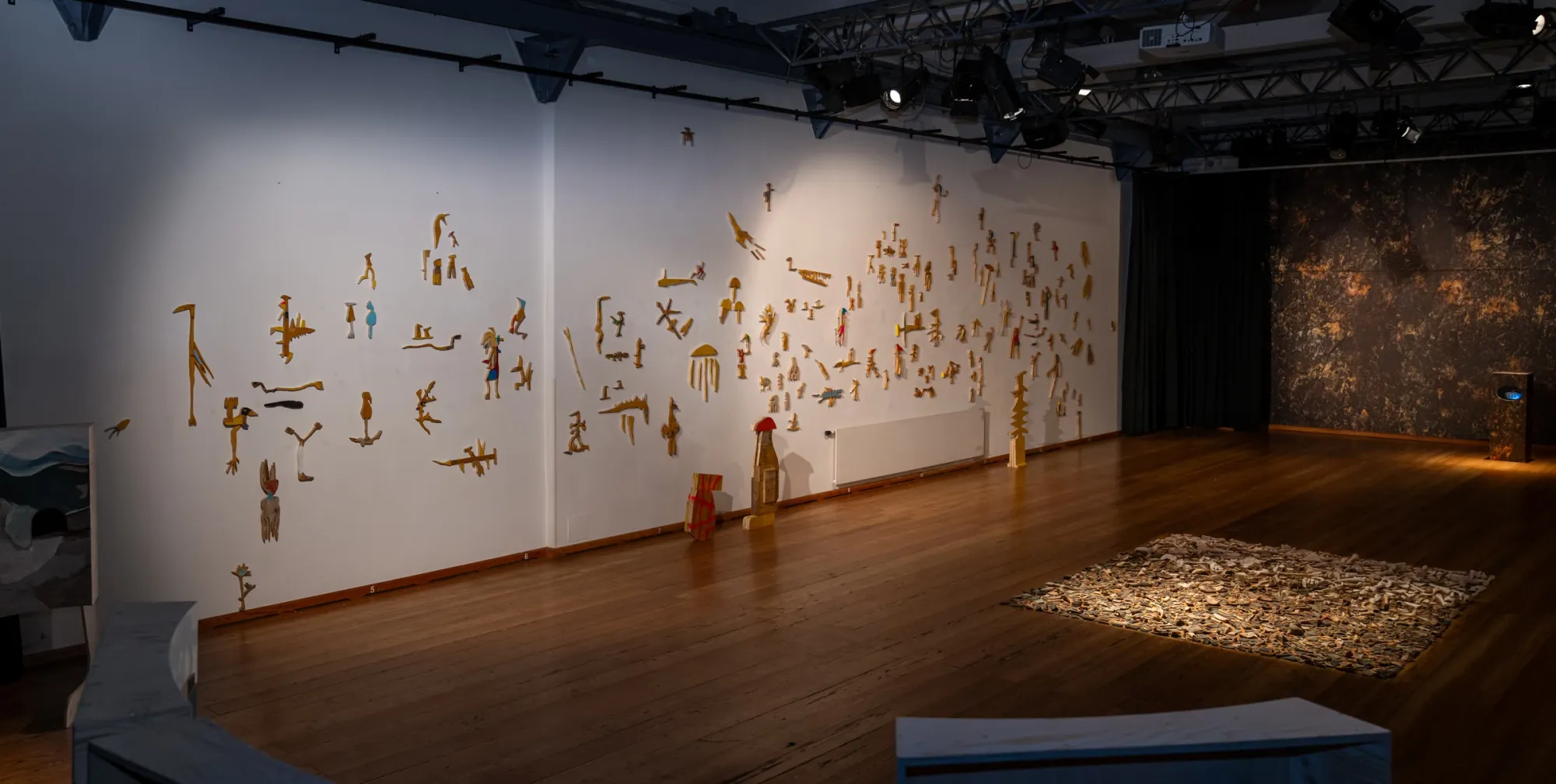 Exhibition review of Ernst Koslitsch: TRANSFER at Kulturforum Laßnitzhaus, Deutschlandsberg, 2026. Three yellow standing wood sculptures positioned in front of the painted wall mural.