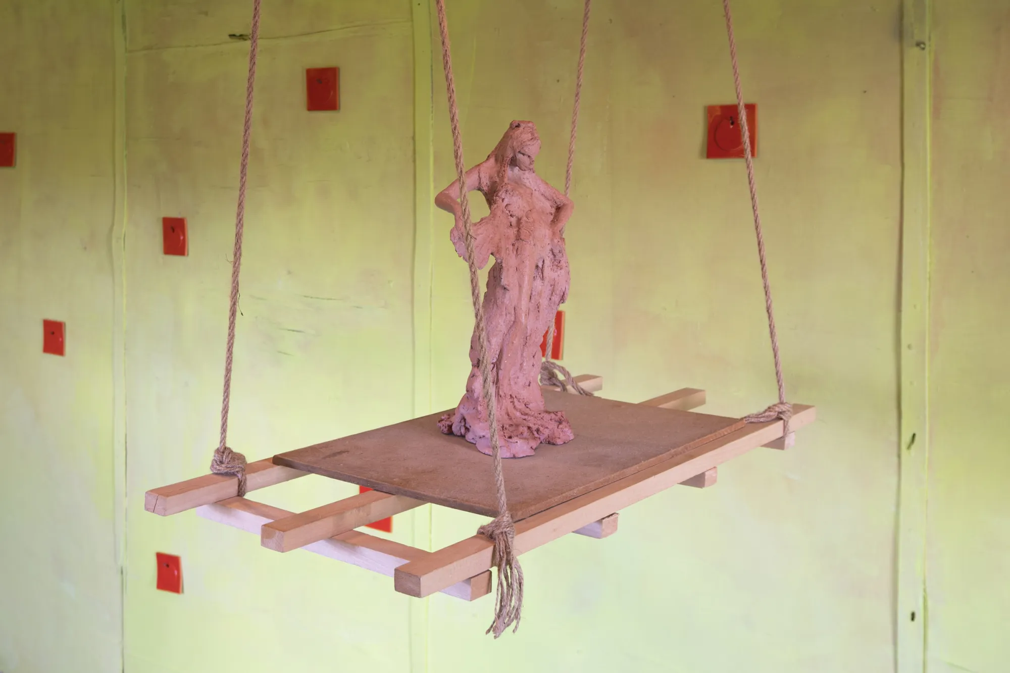 Exhibition review of Tsvetomira Borisova: A Hot Day at lo3eto, Vetren dol, 2026. A red clay female figure standing on a rope-suspended wooden platform.