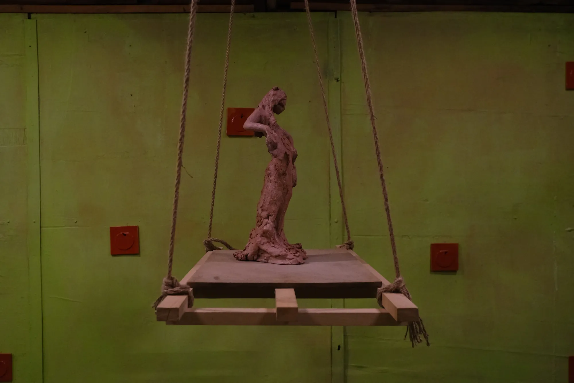 Exhibition review of Tsvetomira Borisova: A Hot Day at lo3eto, Vetren dol, 2026. Side view of the red clay figure on its rope-suspended platform against green walls.