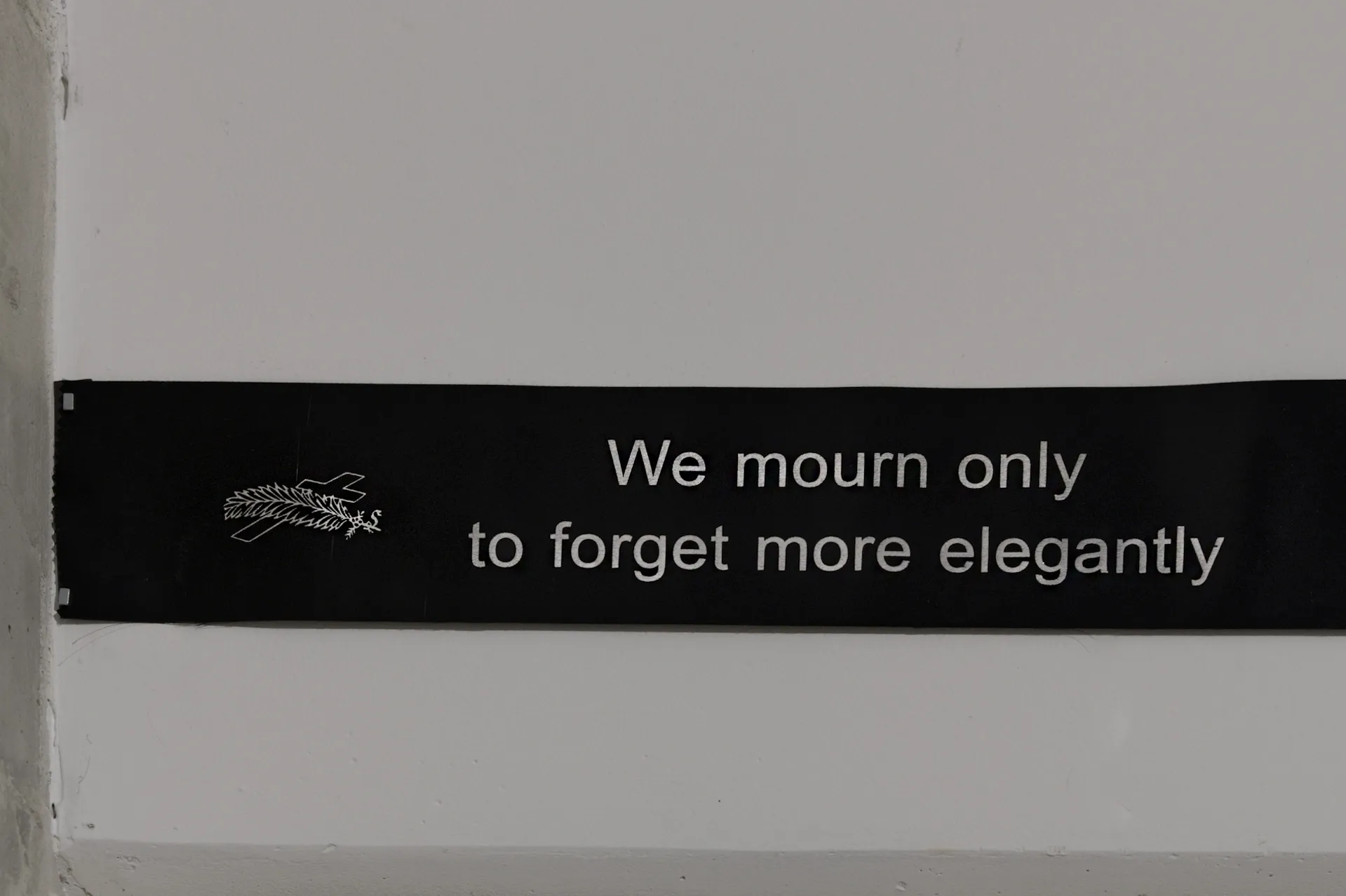 Exhibition review of Sofiia Yesakova: The Solemnity of the Repulsive at ROAM projects e.V., Berlin, 2026. Black funeral ribbon on a white wall with a cross and branch motif and printed text.