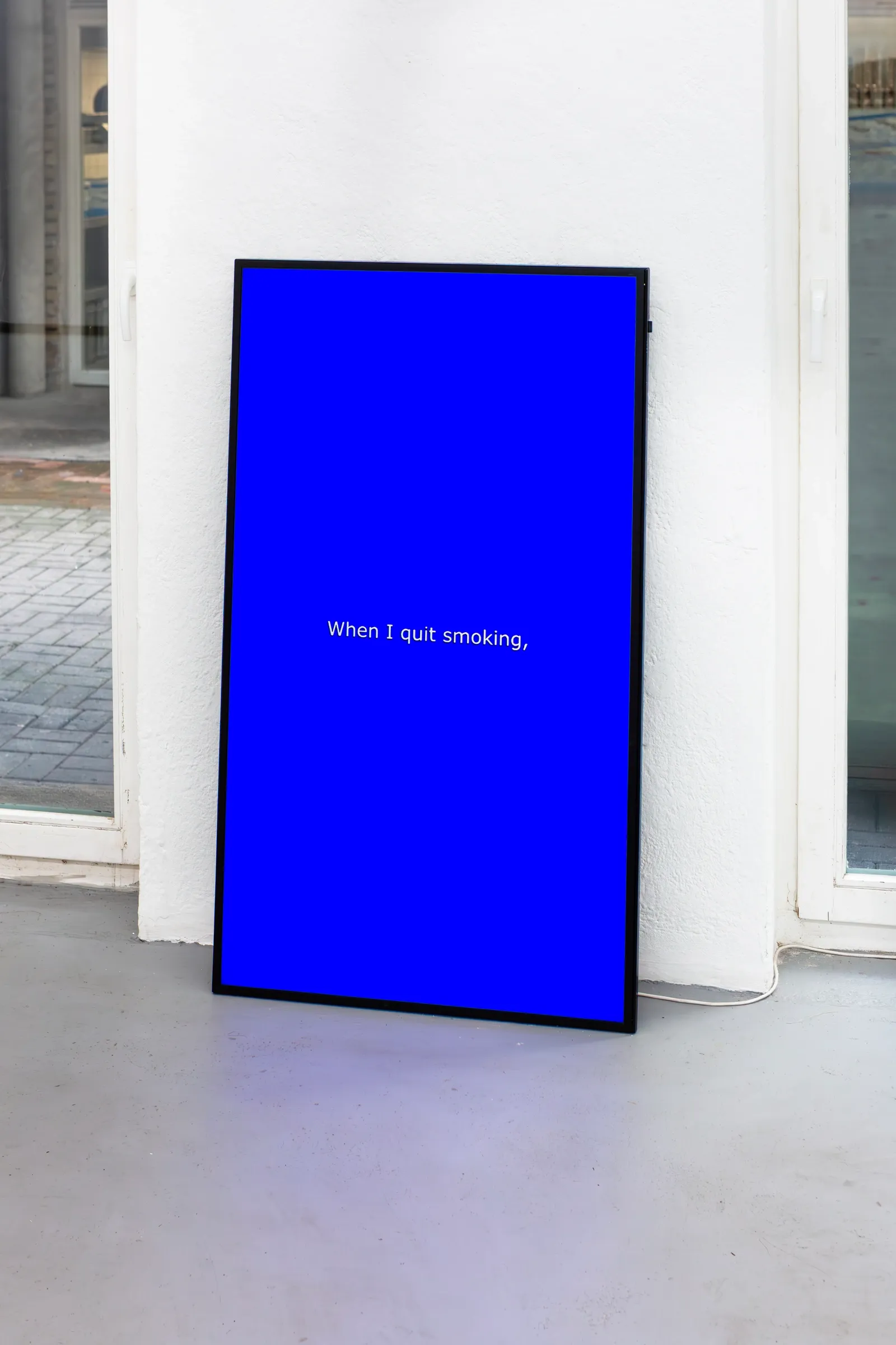 Exhibition review of Anastasia Sosunova: A Charm of Powerful Trouble at ACUD Galerie, Berlin, 2026. A blue video screen leaning against a window frame showing white text reading When I quit smoking.