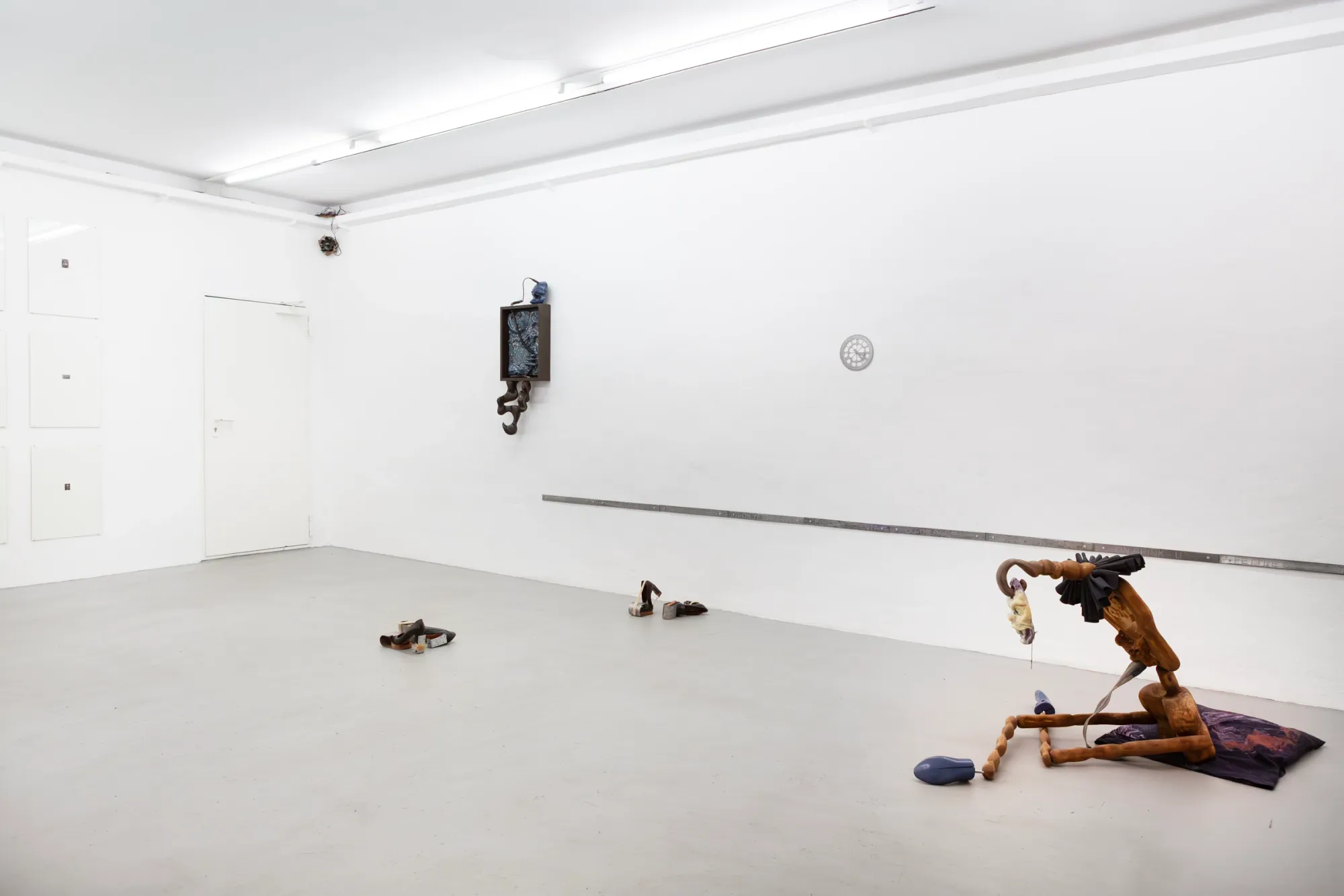 Exhibition review of A Charm of Powerful Trouble at ACUD Galerie, Berlin, 2026. Gallery corner with a decorative wall clock, a white door, and a seated rope-legged sculpture on purple fabric in the foreground.