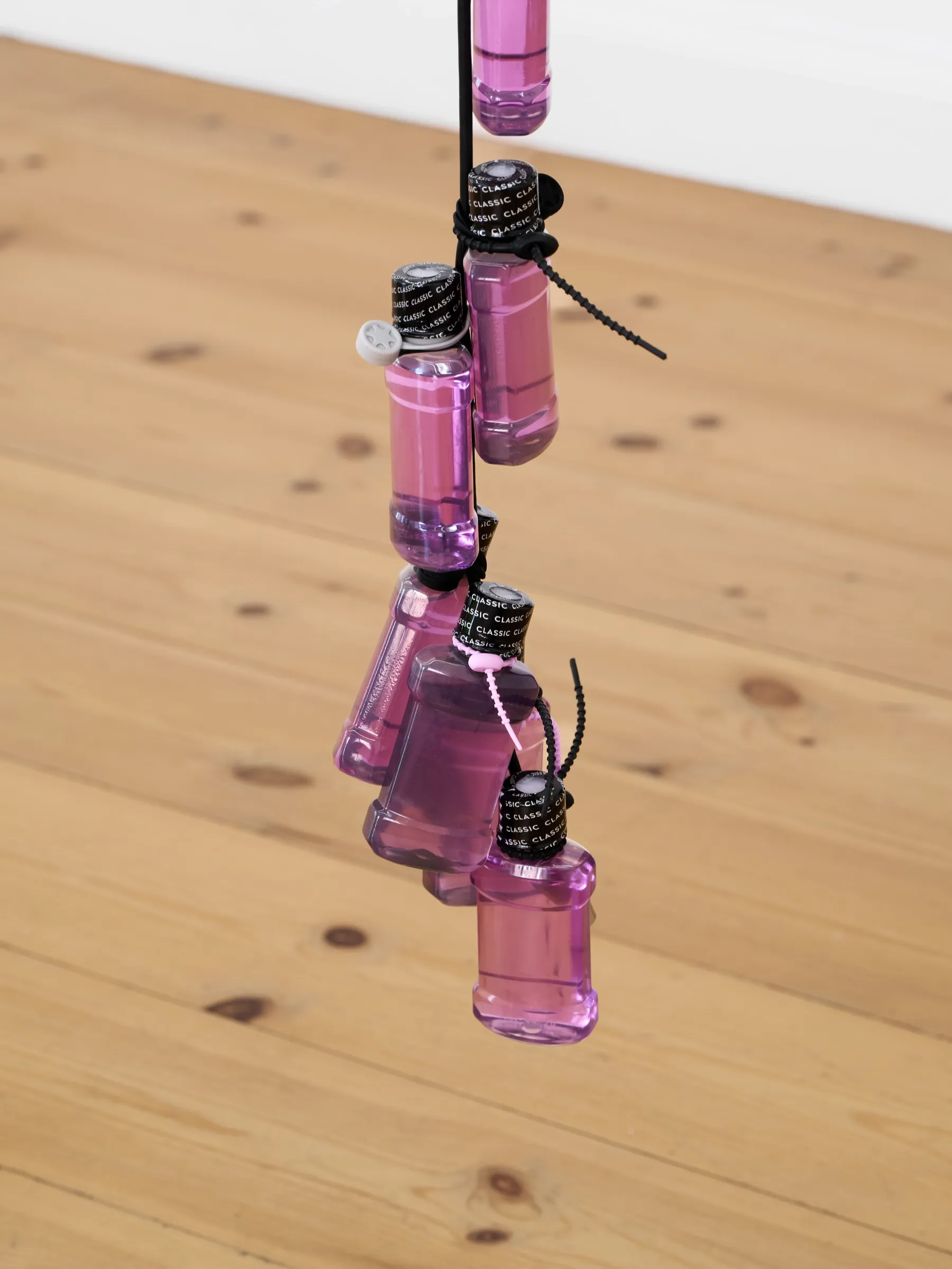 Exhibition review of Elisa Diaferia & Juri Simoncini: Sequence for an Optimal Dispatch at aaaa nordhavn, Copenhagen, 2026. Pink mouthwash bottles tied to a black cord above a pine floor.