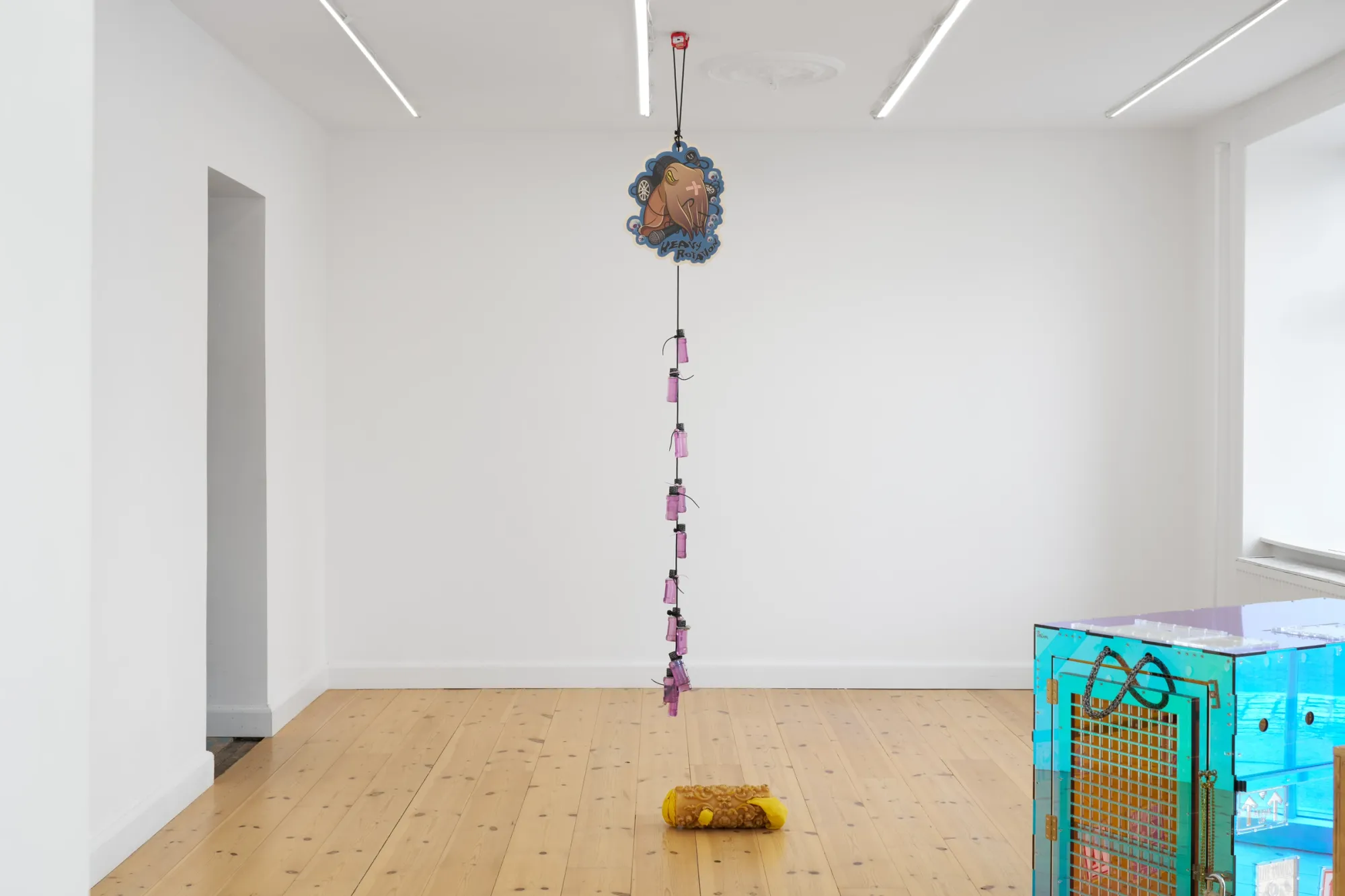 Exhibition review of Elisa Diaferia & Juri Simoncini: Sequence for an Optimal Dispatch at aaaa nordhavn, Copenhagen, 2026. Printed cut-out on a ceiling cord with pink bottles, yellow roll below.