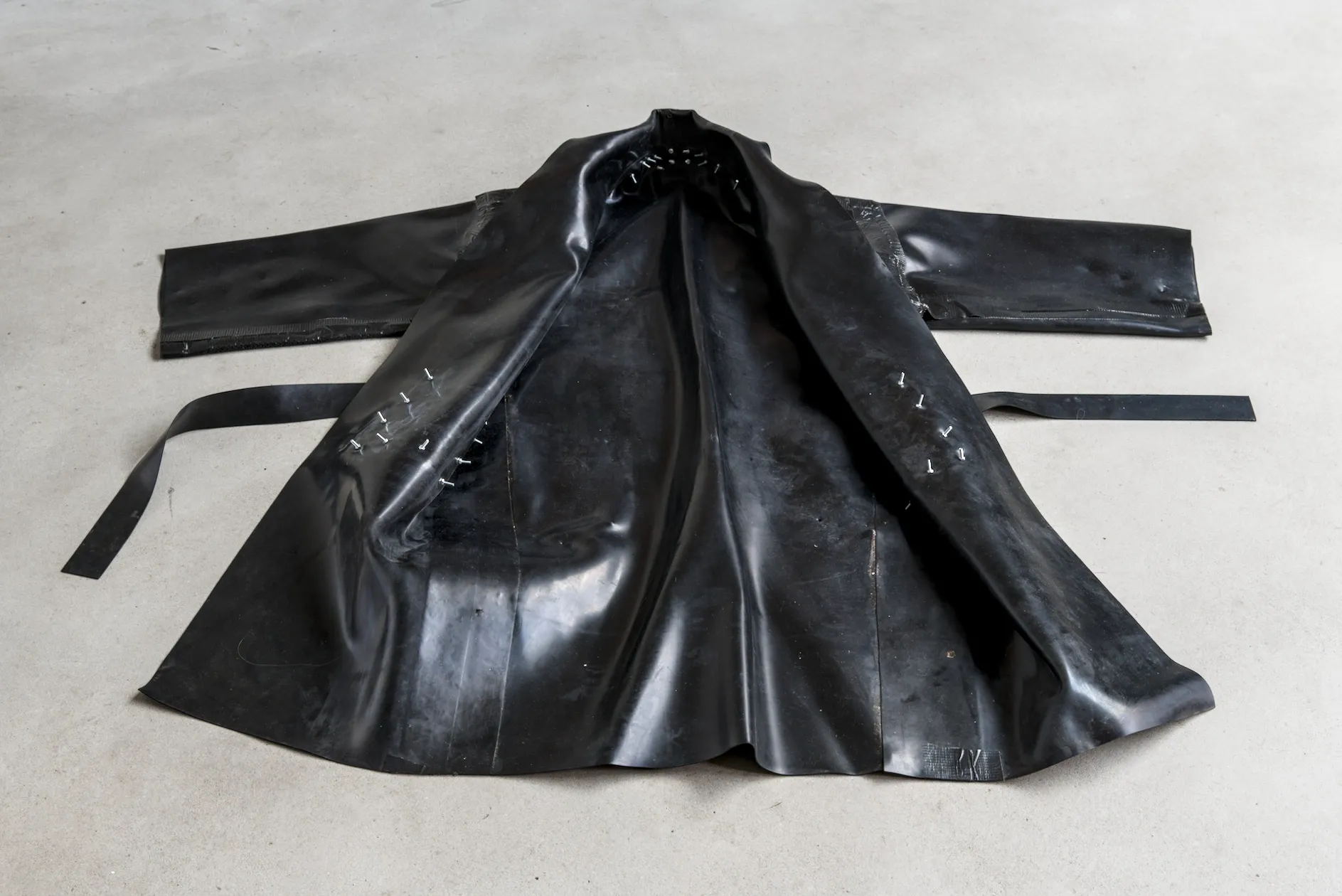Exhibition review of I Think My Body No Longer Belongs to Me at roam projects e. V., Berlin, 2026. Black rubber coat laid open on grey floor, bolted seams.