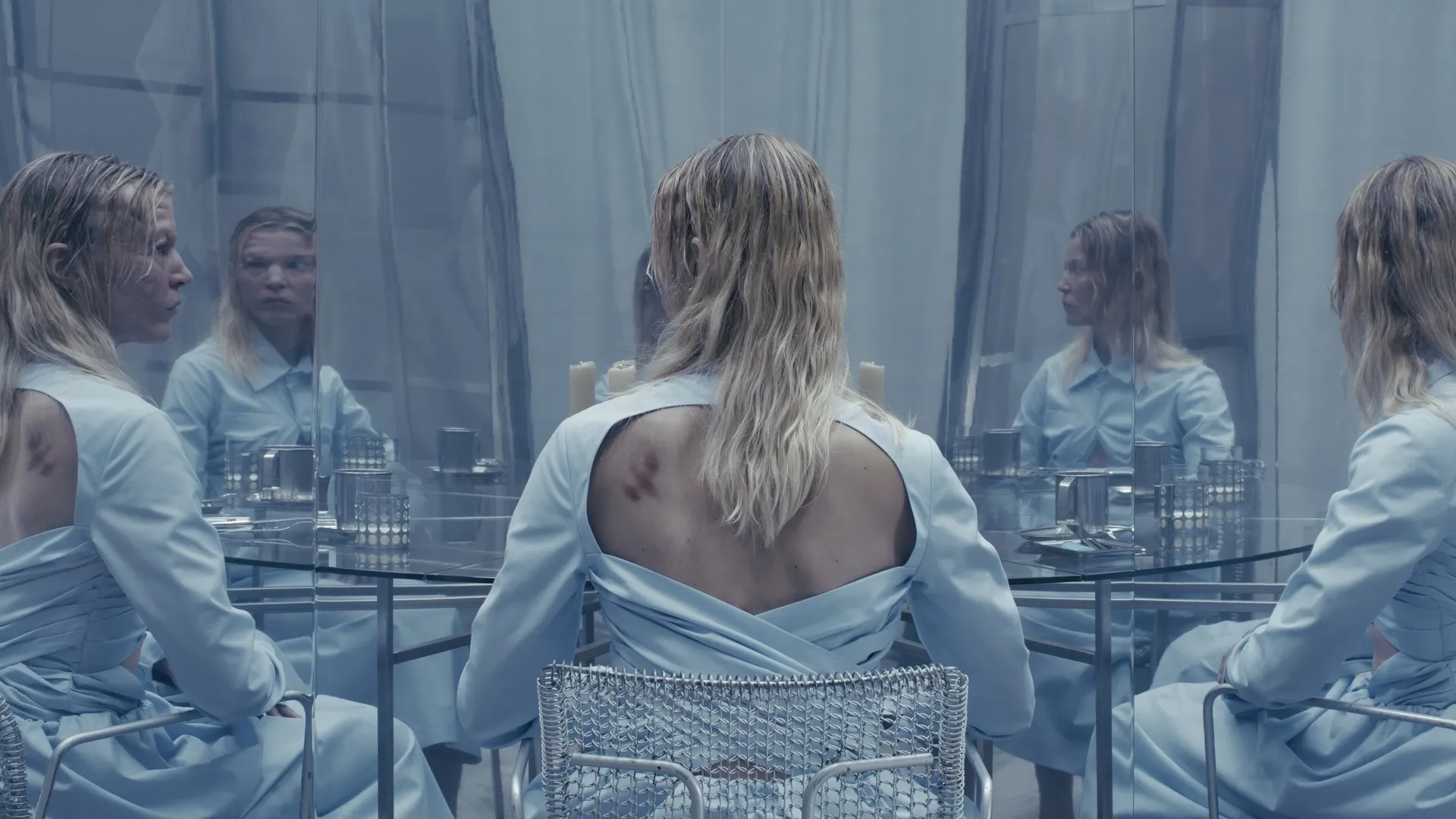 Exhibition review of I Think My Body No Longer Belongs to Me at roam projects e. V., Berlin, 2026. Blonde women in pale blue at a mirrored glass table