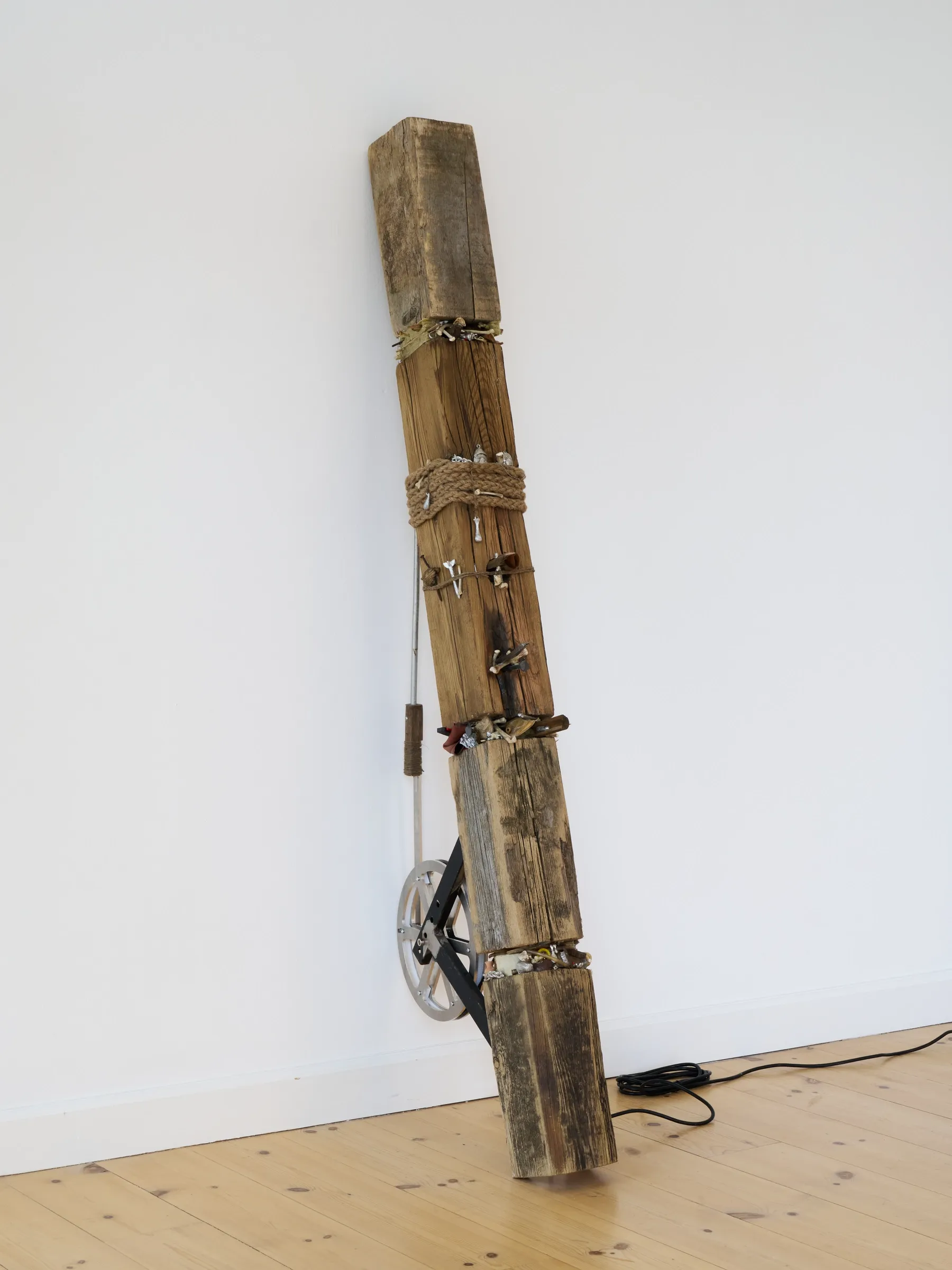 Exhibition review of Gustav Gaston: Sequence for an Optimal Dispatch at aaaa nordhavn, Copenhagen, 2026. Weathered driftwood post bound with rope and objects, metal wheel at its base.